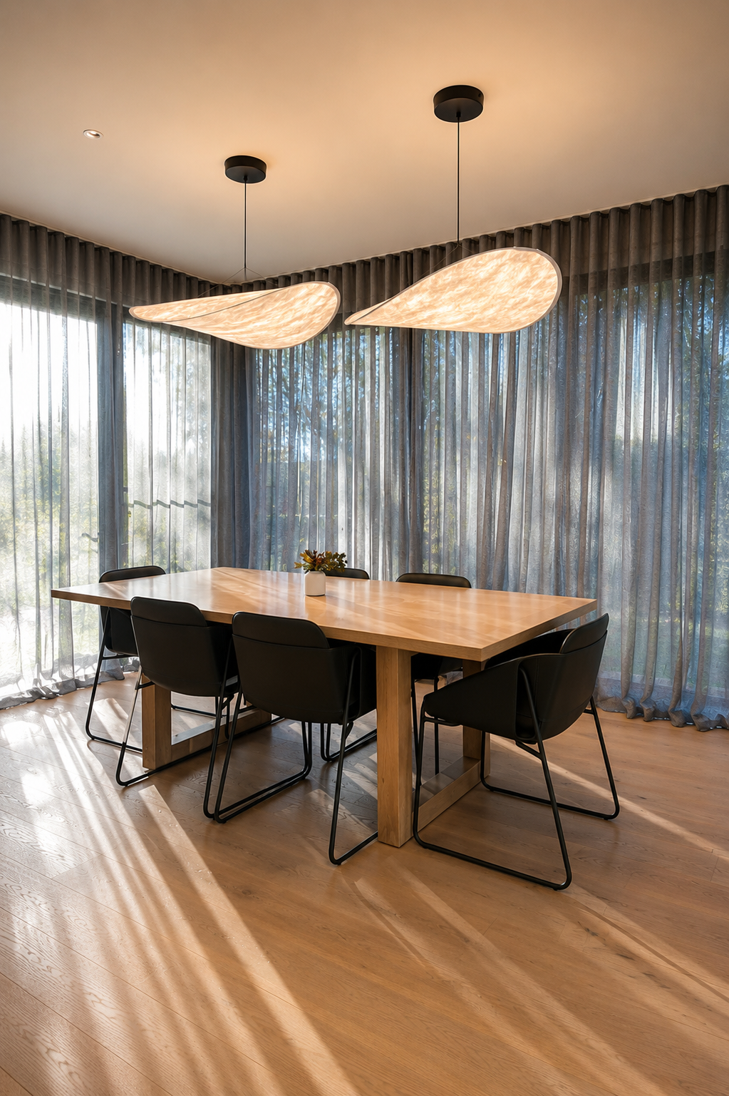 A dining room with a wooden table surrounded by six black chairs, featuring two modern hanging ceiling lights and large windows with sheer curtains, allowing natural light to fill the space.