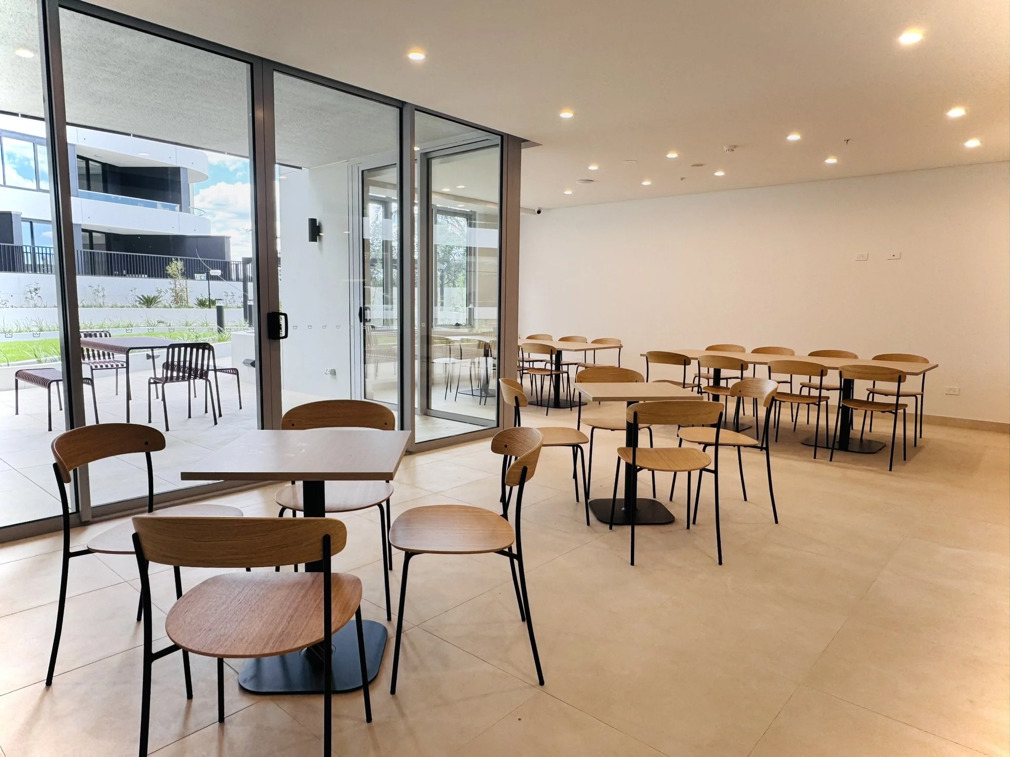 Empty modern cafeteria with wooden chairs and tables, large glass doors leading to an outdoor patio.