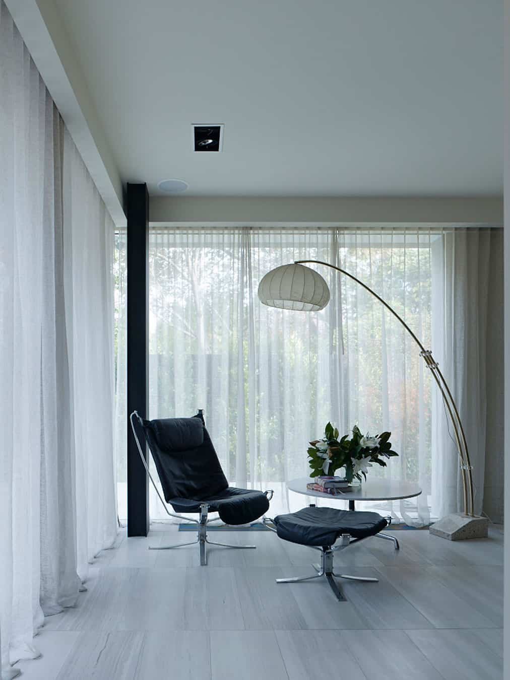 Modern living room with a black leather lounge chair and matching ottoman, a white round side table with books and a plant, a tall arc floor lamp, sheer white curtains, and large windows showing trees outside.