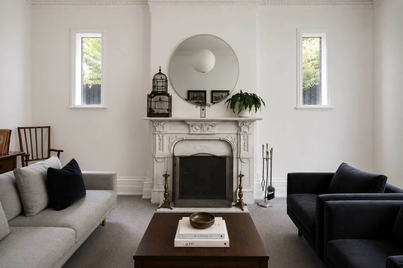 Living room with a white ornate fireplace, a large round mirror above it, and two small windows on each side. Decor items on the mantle include a birdcage, framed pictures, and a potted plant. There are two sofas: a light-colored one on the left with
