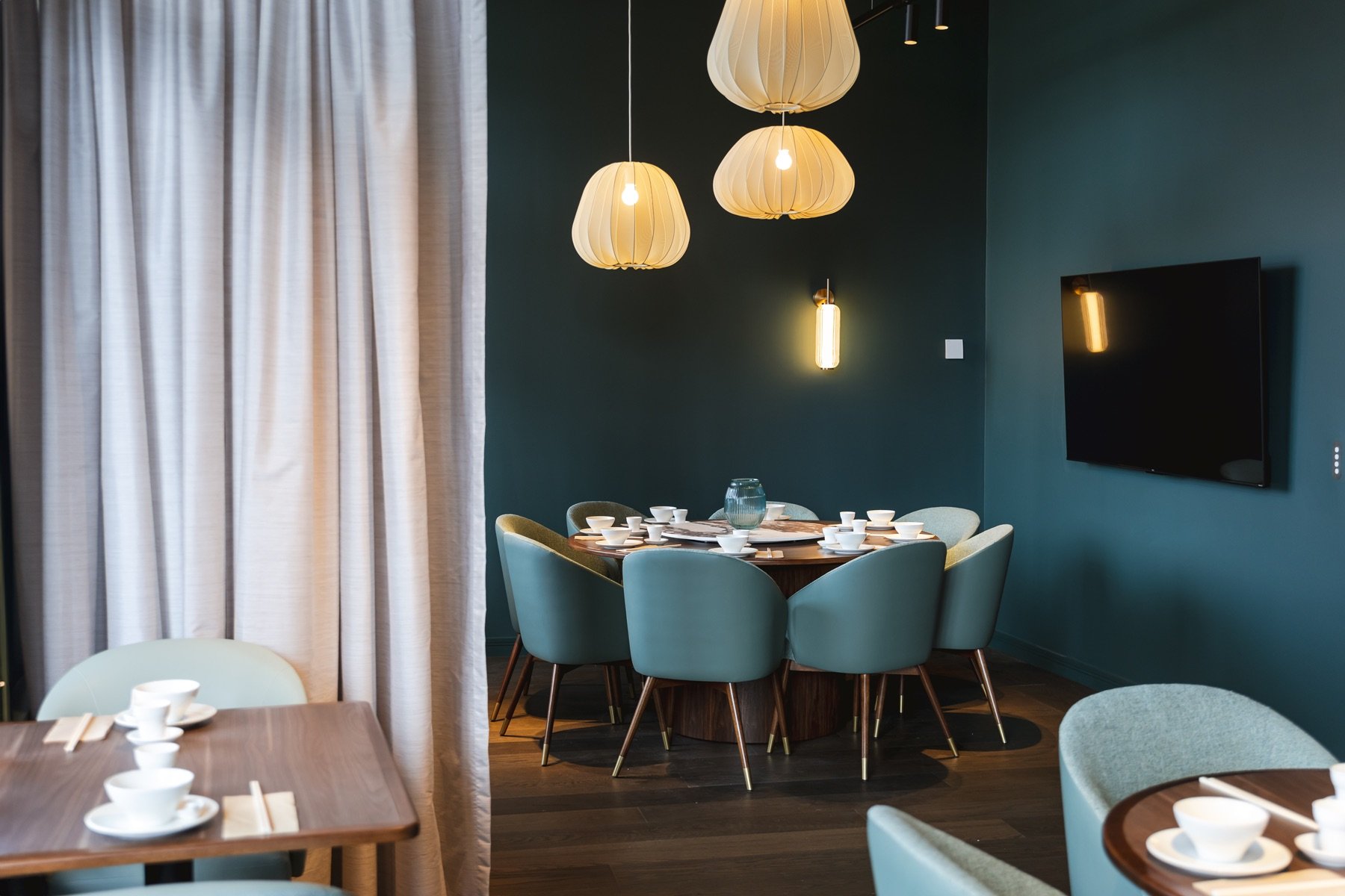 A dining room with teal walls, a round wooden table set with white bowls and chopsticks, surrounded by teal chairs, cream curtains, and modern hanging lights. A flat-screen TV is mounted on the wall.