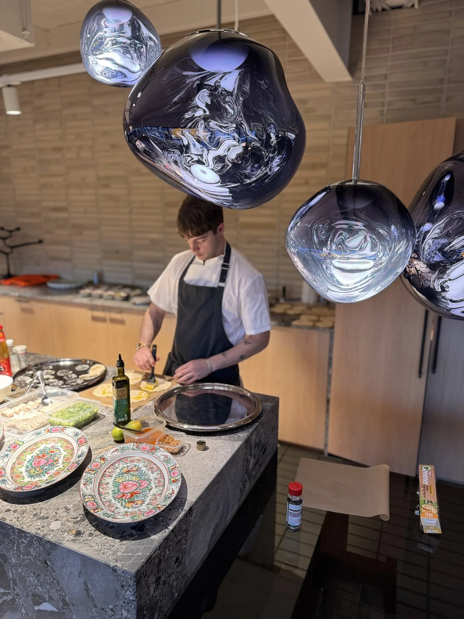 A man in a white shirt and black apron preparing food on a kitchen counter with decorative plates, ingredients, and condiments, with modern hanging glass pendant lights overhead.