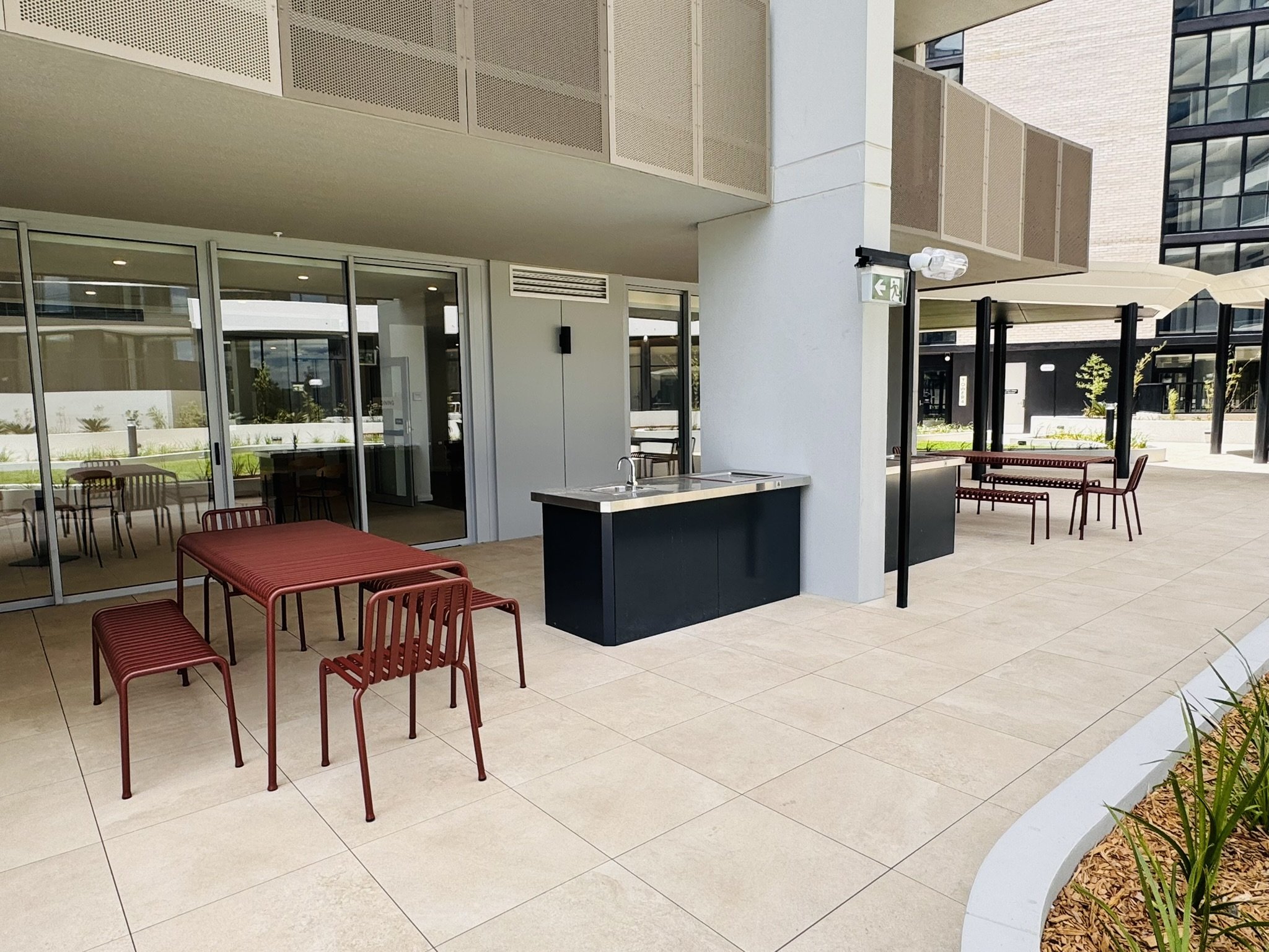 Outdoor patio with red metal chairs and tables, built-in sink, glass doors, and umbrellas in a modern residential or commercial building complex.