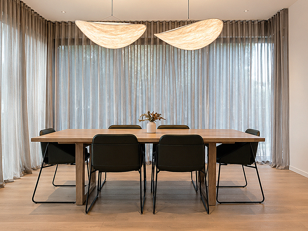 Modern dining room with a wooden table, six black chairs, sheer curtains, and two large modern ceiling lights.
