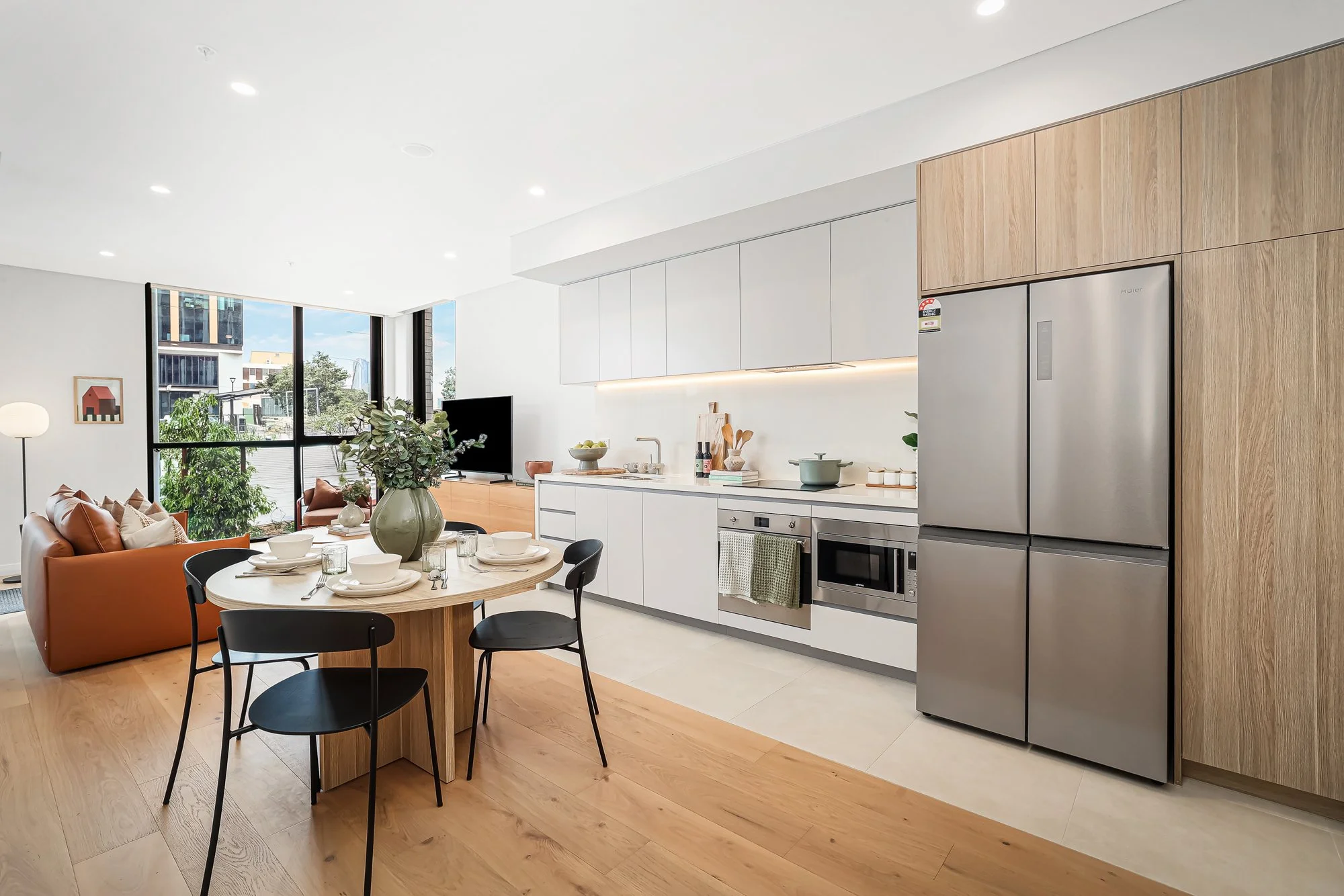 Open-concept modern kitchen and living area with wooden flooring, white cabinetry, stainless steel appliances, a dining table with black chairs, and a balcony with outdoor furniture and city view.