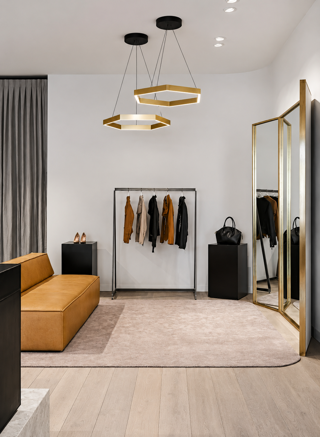 A modern boutique interior with a mustard-colored sofa, black side tables, a clothing rack with beige, black, and brown jackets, a black handbag on a pedestal, gold-framed full-length mirror, and stylish gold geometric pendant lights hanging from the ceiling.