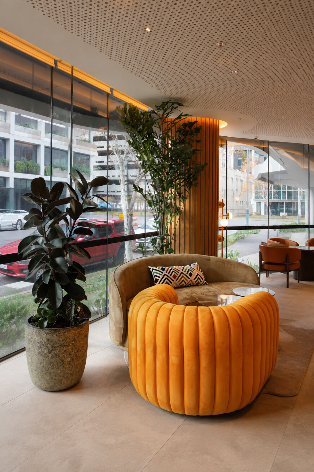 Modern hotel lobby with large glass windows, green plants, and contemporary seating including a curved beige sofa with a colorful patterned pillow and an orange velvet chair in front of a window.