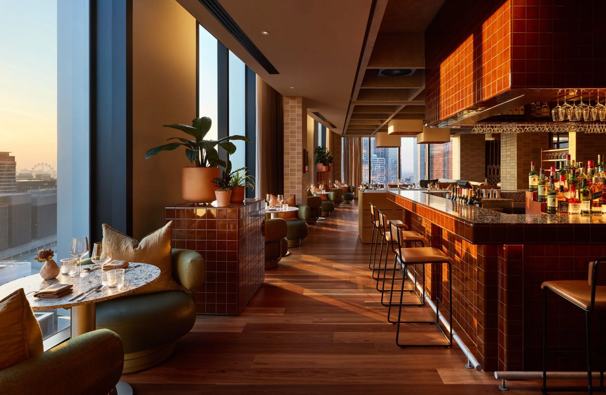 Modern restaurant interior with large windows showing a cityscape, a bar area with bottles of liquor, and elegant seating with tables set for dining.
