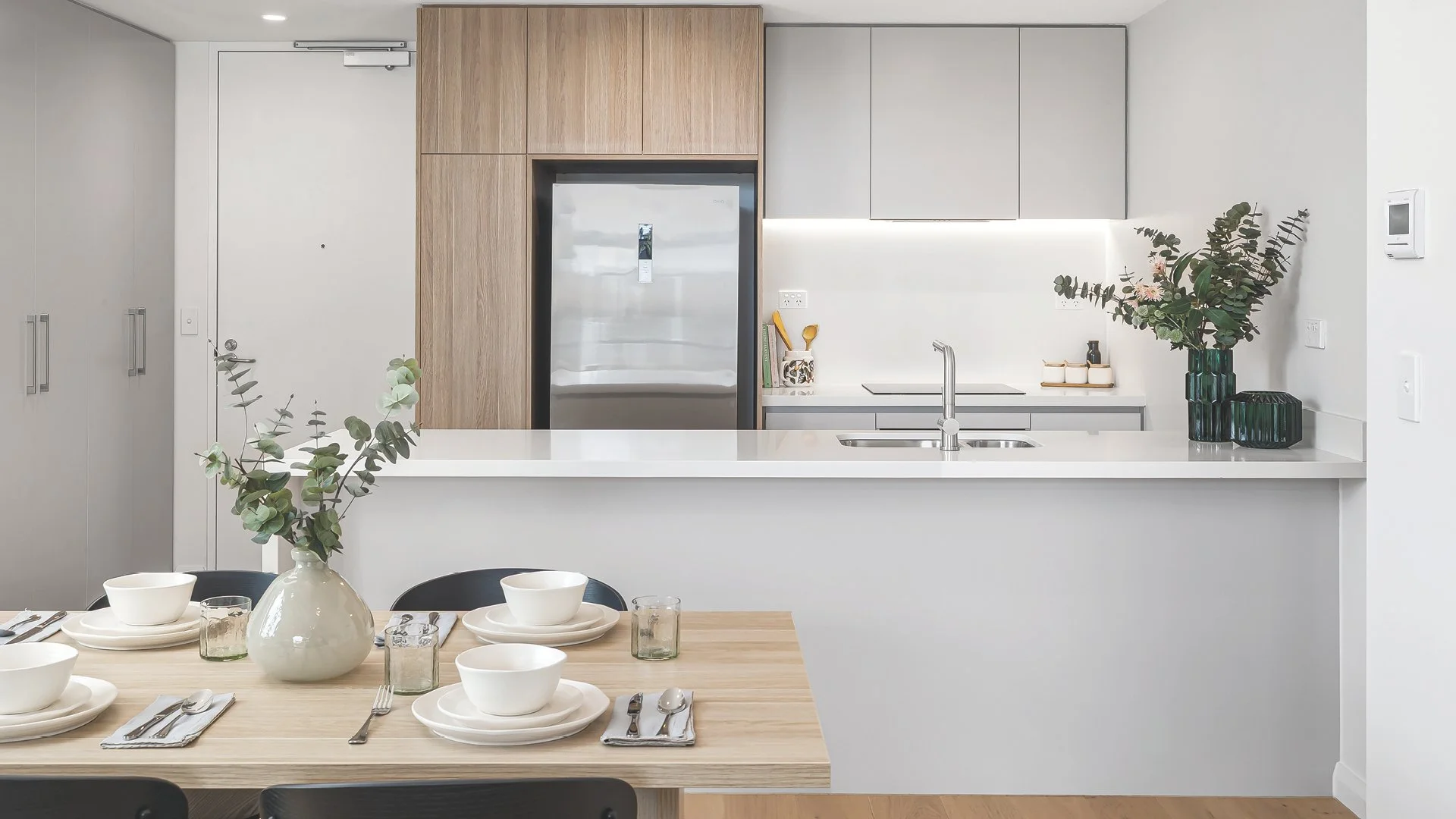 Modern kitchen with white countertops, gray and wooden cabinets, a stainless steel refrigerator, and a dining table set with bowls, plates, glasses, and a vase with green foliage.
