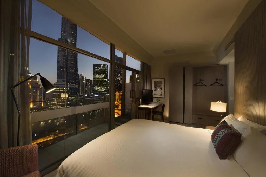 A modern hotel room with a large bed, a balcony with city views, a desk with a chair, and a closet with hanging space, illuminated by a bedside lamp at dusk.