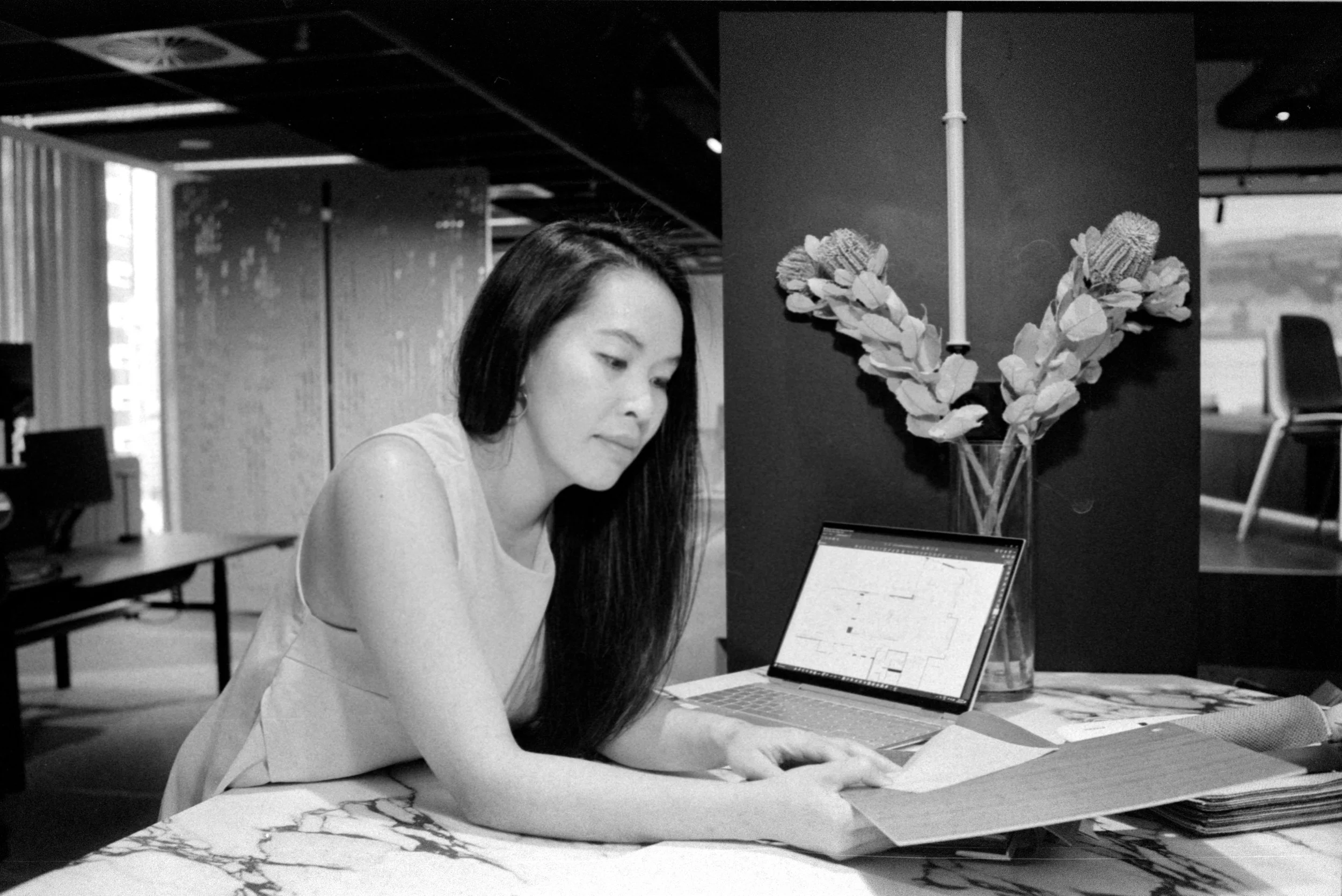 A woman with long dark hair working at a marble table in a modern office, looking through papers with an open laptop nearby and a vase with flowers behind her.