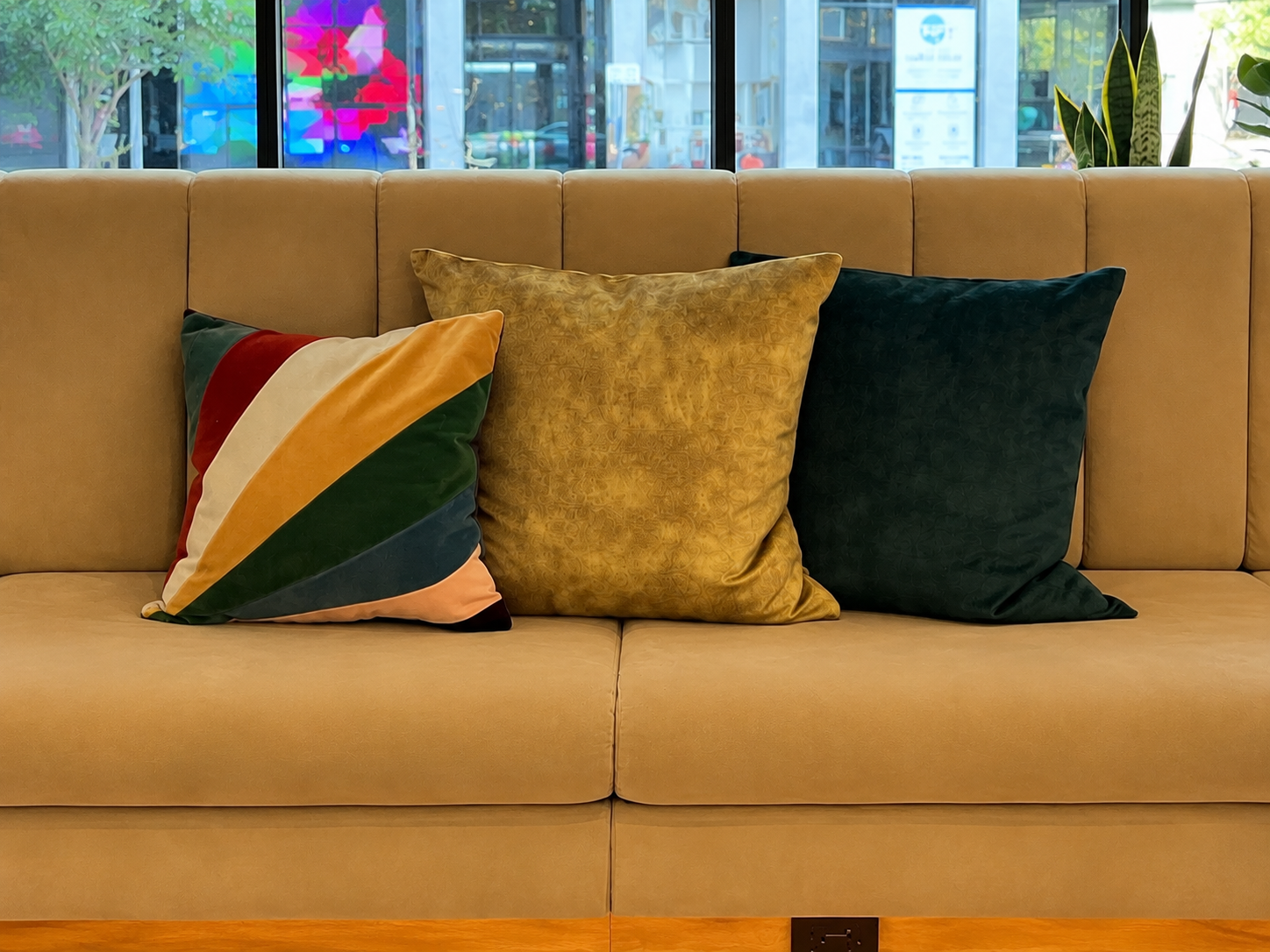 A beige sofa with three decorative pillows: a multicolored striped pillow, a gold pillow, and a dark green pillow. A window behind the sofa shows a city street scene with storefronts, signs, and street view.