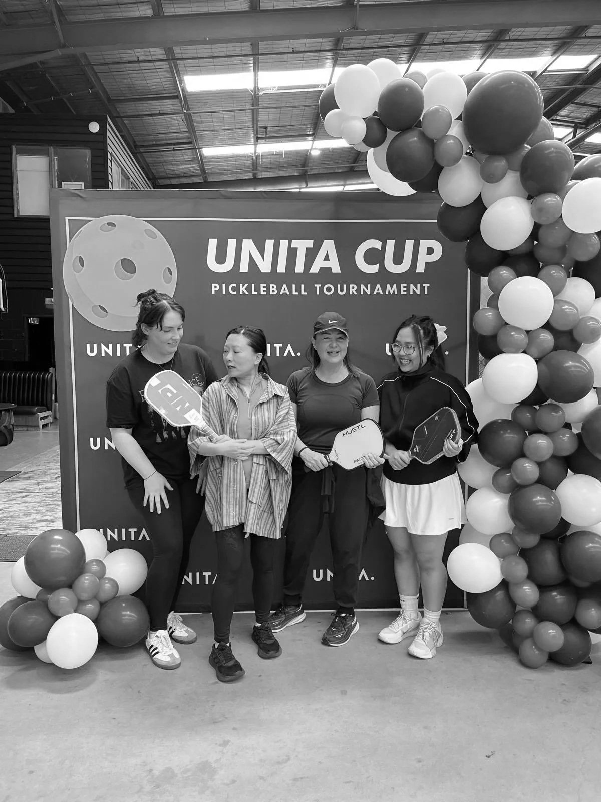 Four women standing in front of a backdrop with balloons at the UNITA Cup Pickleball Tournament, holding pickleball paddles and smiling.