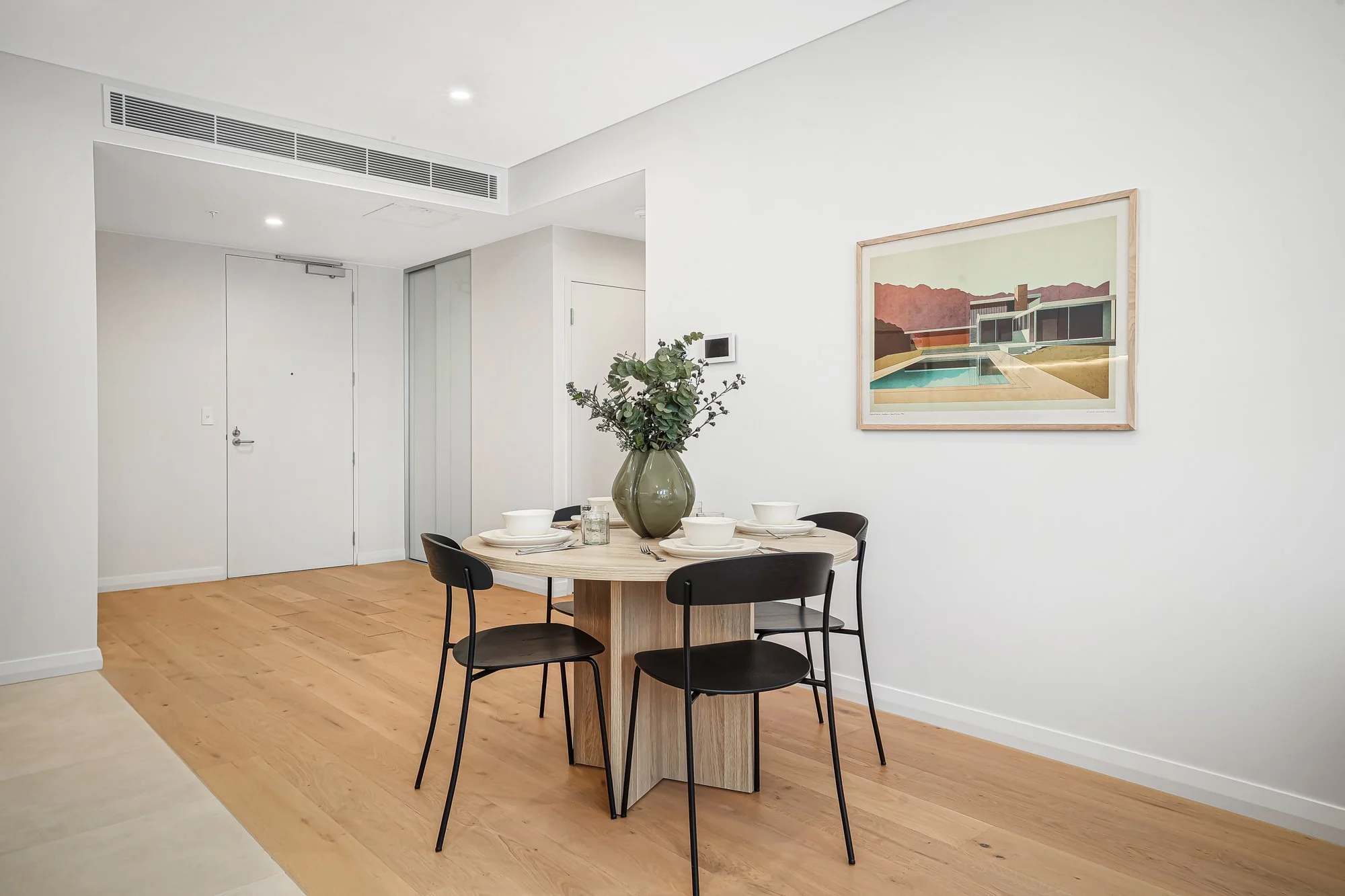 Dining area with a wooden table, black chairs, a large green vase with greenery, and a framed landscape artwork on a white wall.
