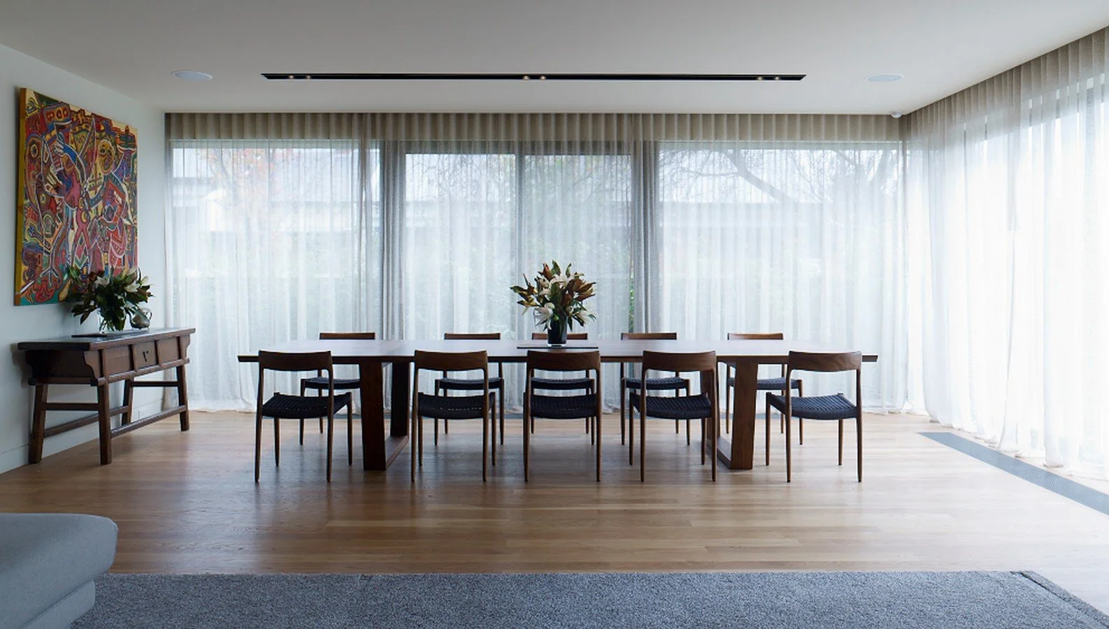 A modern dining room with a long wooden table, eight matching chairs, a window with sheer white curtains, a floral centerpiece, an abstract colorful painting on the wall, and hardwood flooring.