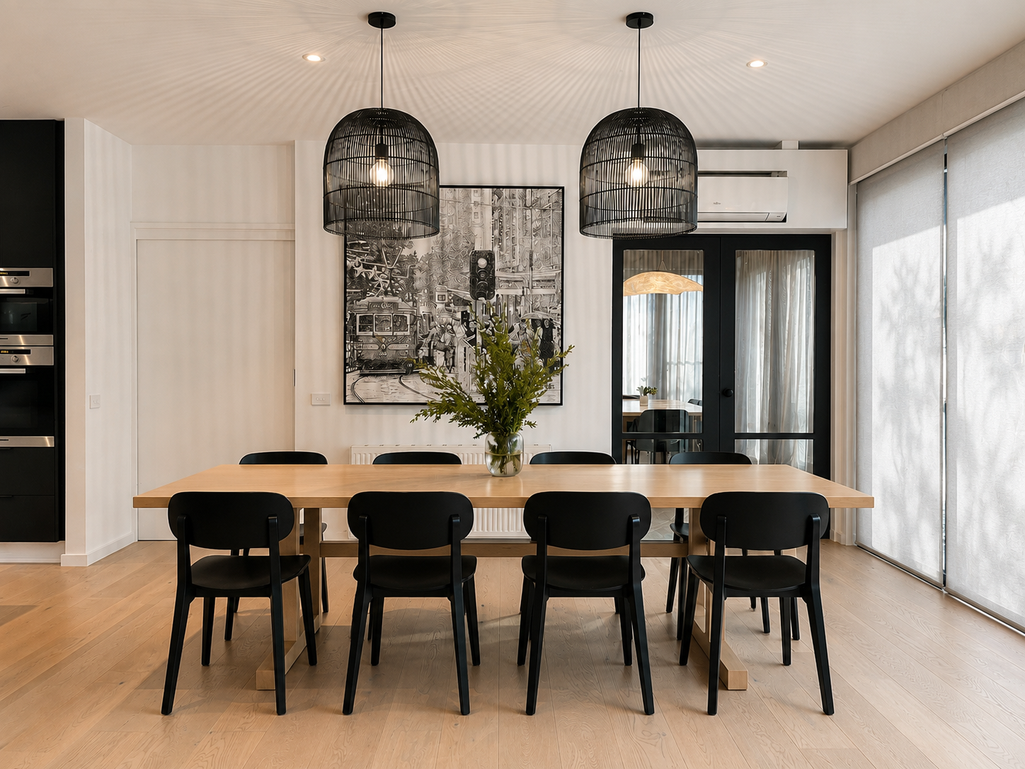 Modern dining room with a wooden table, eight black chairs, a vase with greenery, framed artwork, two black pendant lights, and sliding glass doors with curtains.