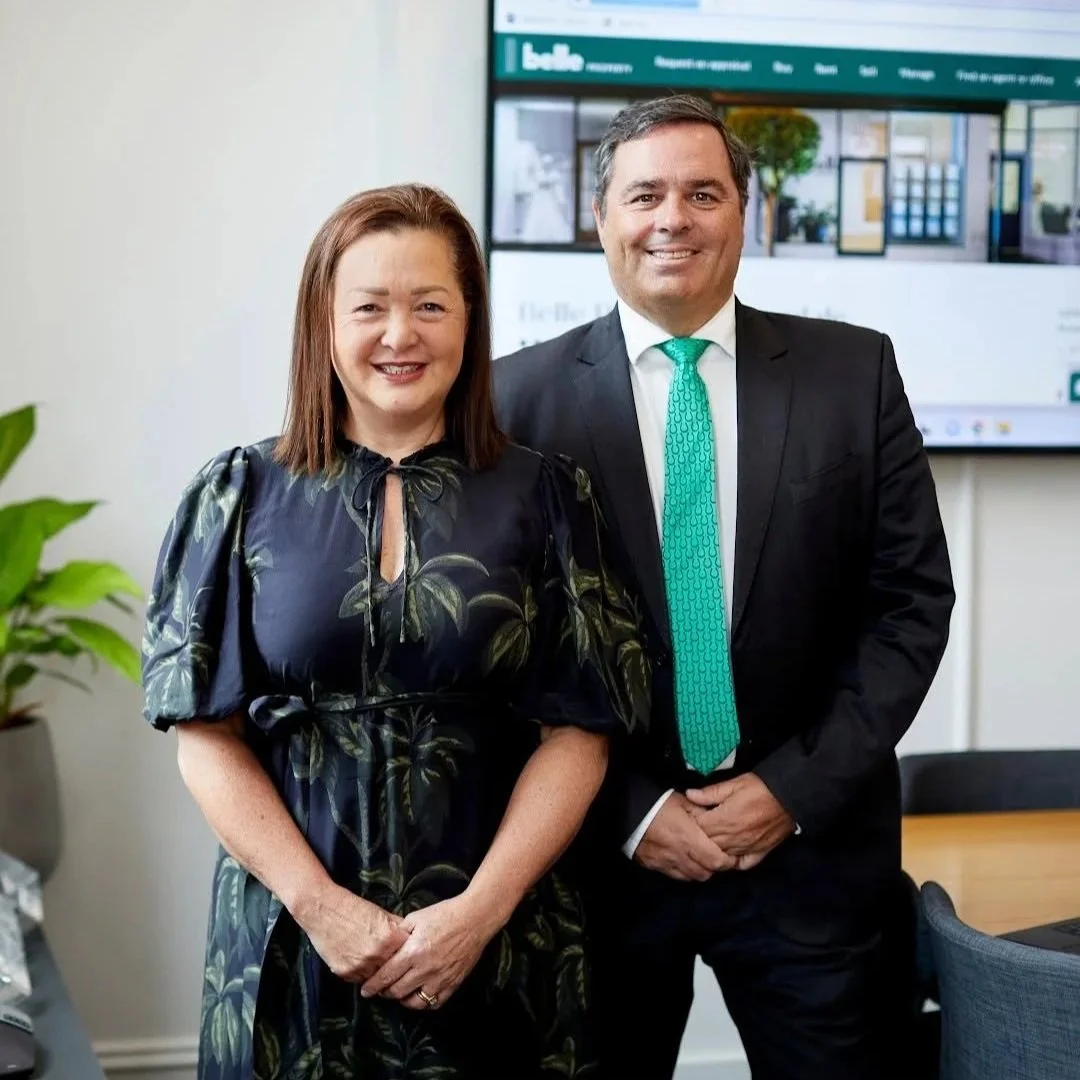 A woman and a man standing side by side, smiling, in a professional setting with a large monitor displaying a website in the background.