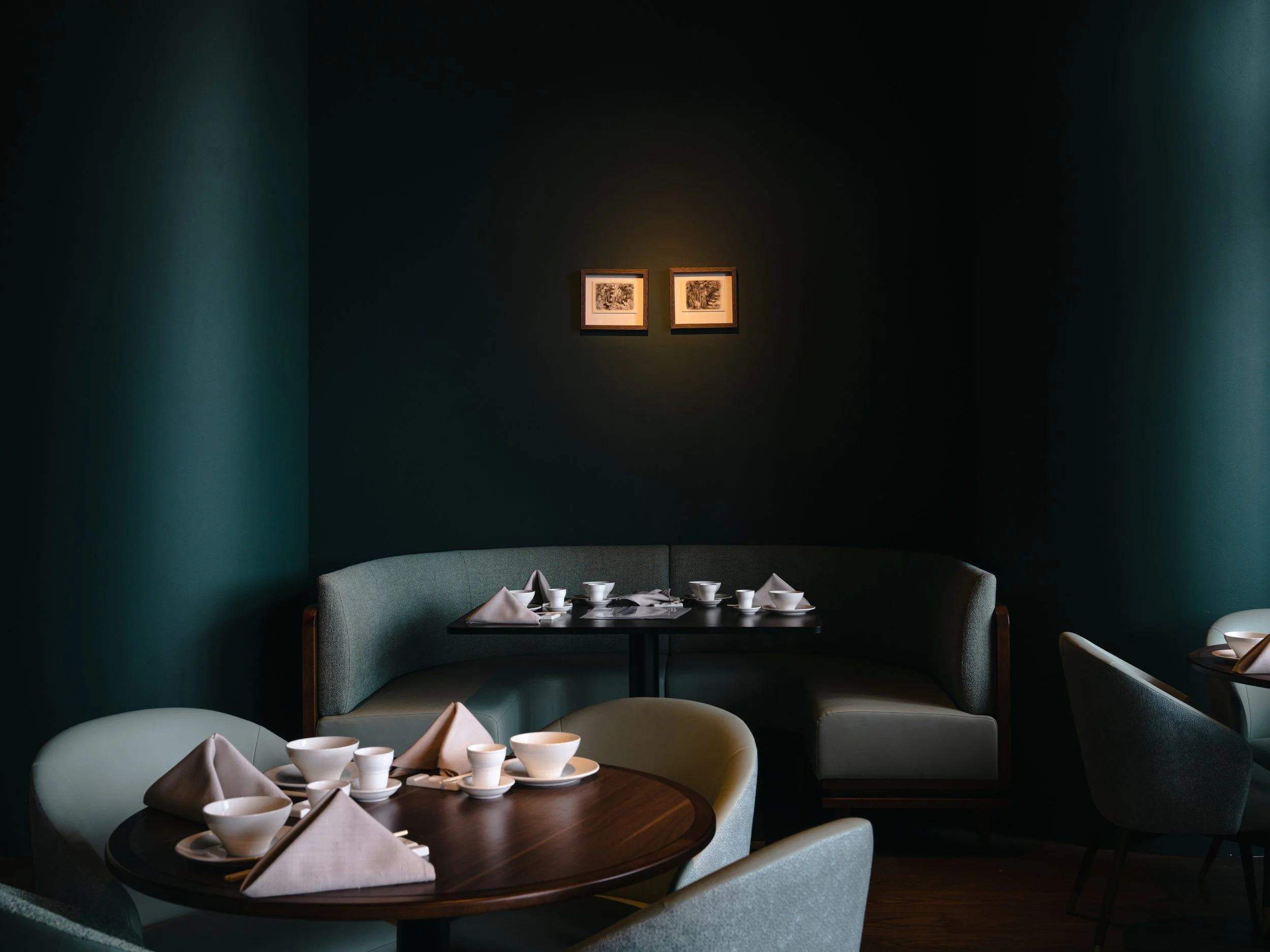 A cozy dining area with a dark circular table set with white bowls, cups, and folded napkins, surrounded by plush, light-colored chairs and a curved booth, with framed artwork on a dark green wall in the background.