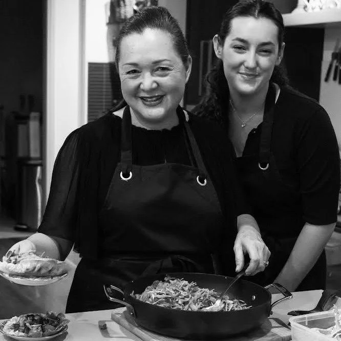 Two women in a kitchen, wearing black aprons, smiling and cooking together, with a frying pan and bowls of ingredients.