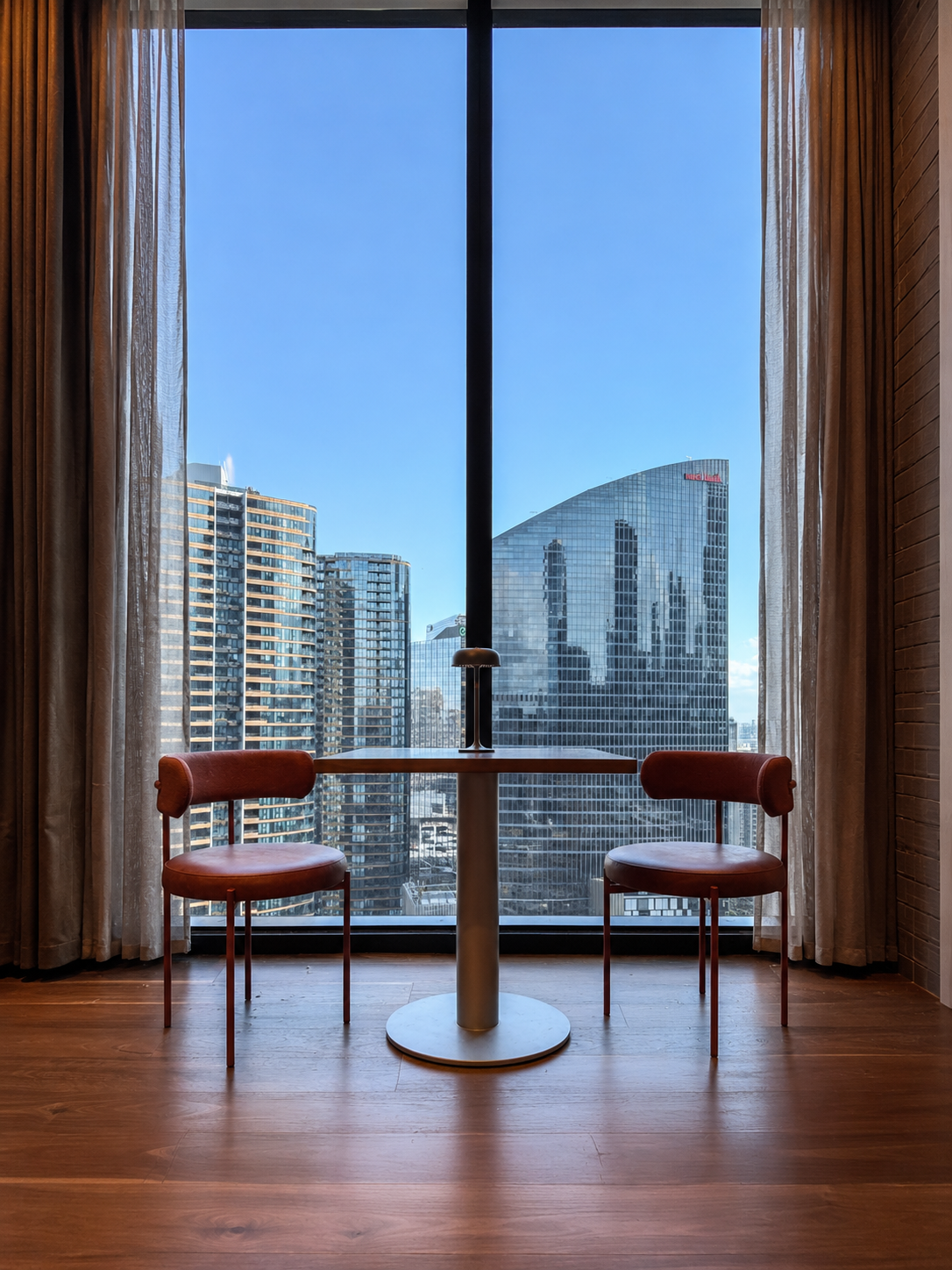 A small round table with a lamp on it, flanked by two red cushioned chairs, positioned in front of large floor-to-ceiling windows with sheer curtains, overlooking a cityscape of modern high-rise buildings against a blue sky.