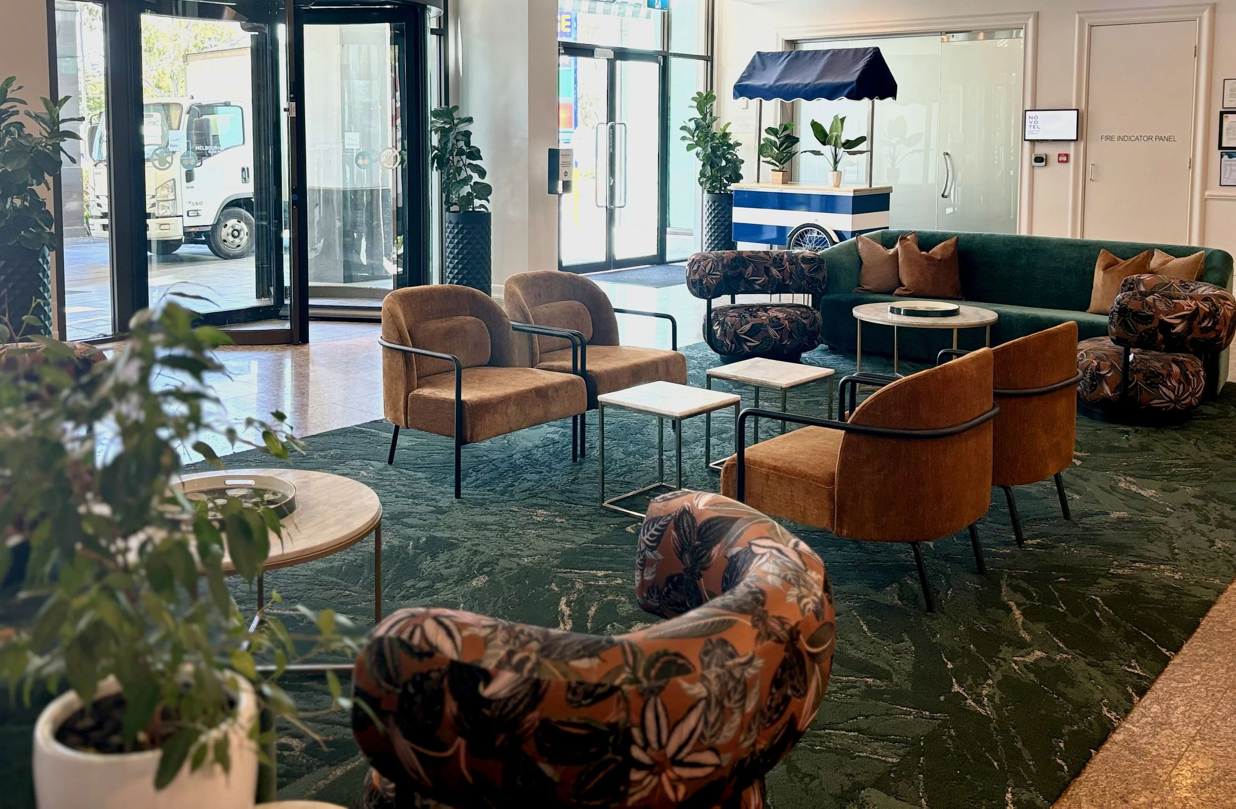 Hotel lobby with modern seating, including brown, green, and floral armchairs, small tables, potted plants, and a glass entrance door.