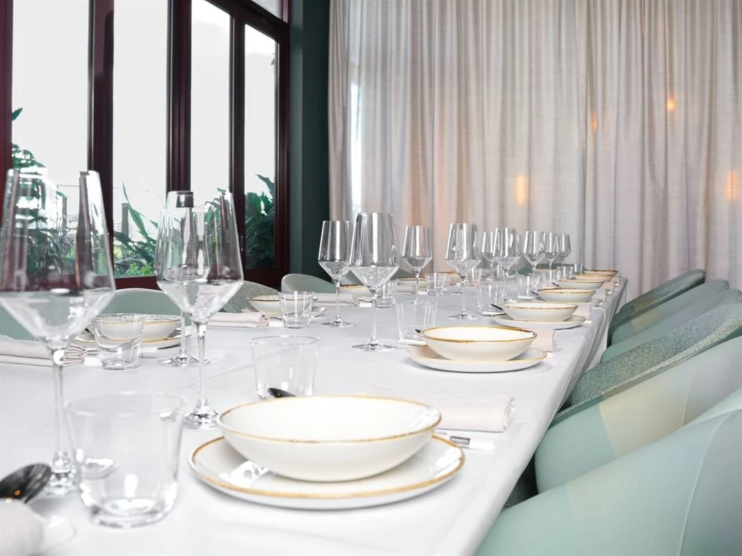 Elegant dining table set with white tablecloth, multiple wine glasses, water glasses, bowls, plates, and silverware, near large windows with white curtains.