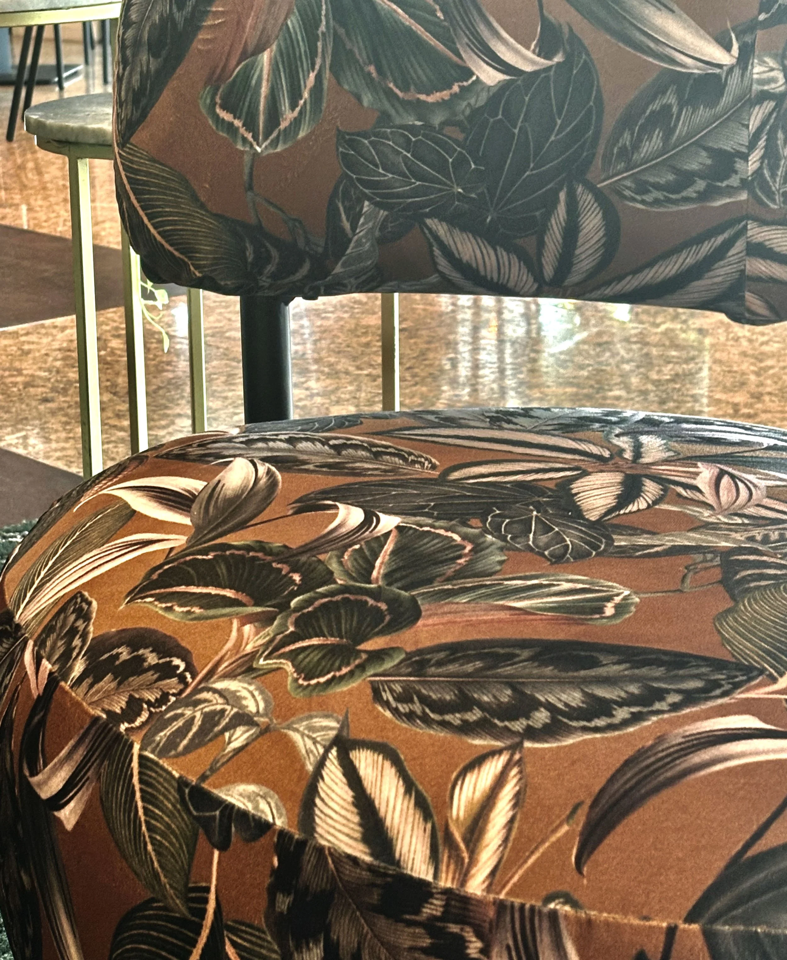 Close-up of two chairs with floral-patterned upholstery, one in the foreground and one in the background, in an indoor setting, possibly a restaurant or cafe.
