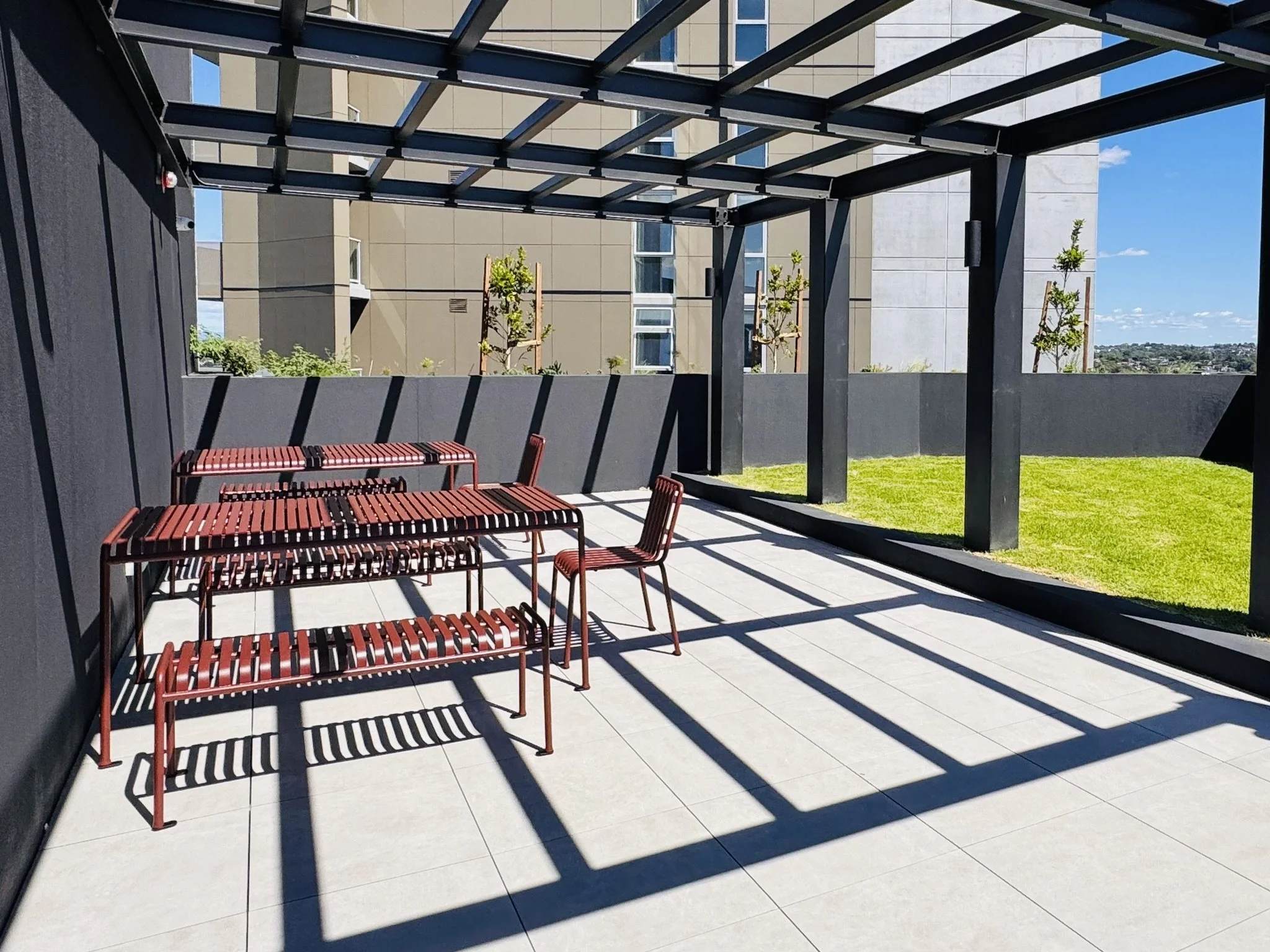 Outdoor terrace with metal tables and chairs, surrounded by a black wall and open on two sides revealing a grassy area and neighboring buildings. Shadows of the framework cast on the light-colored tiled floor, with a bright blue sky in the background