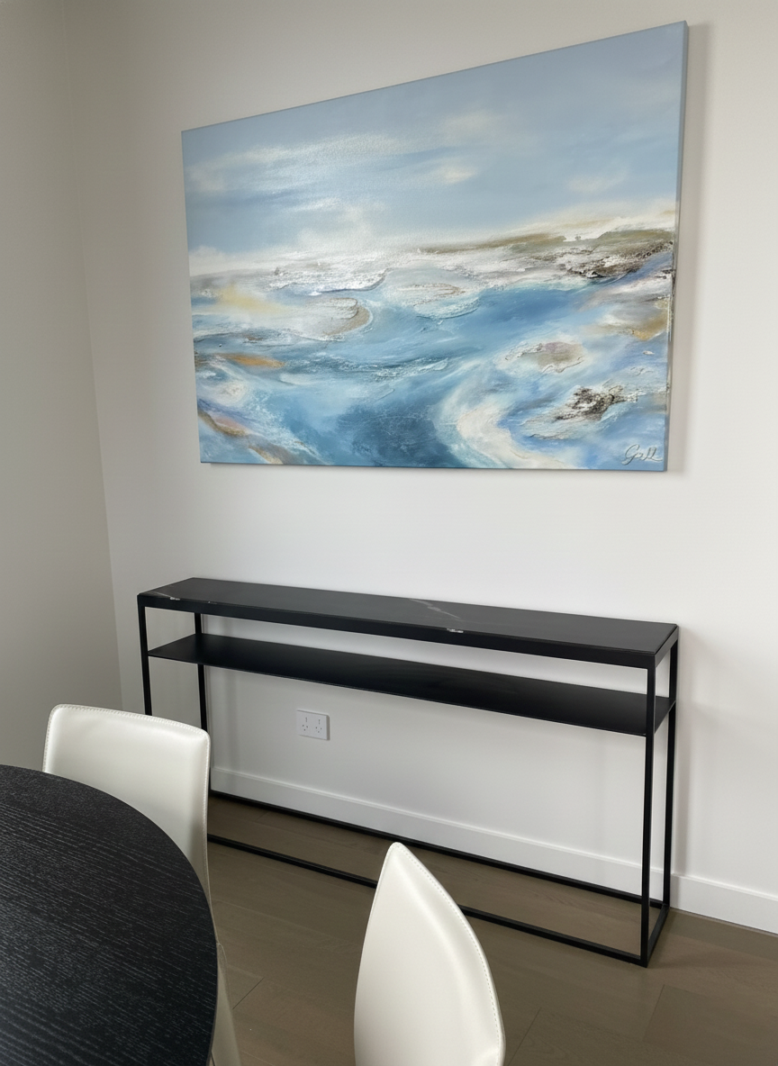 A contemporary living room or dining area with a white wall, a large abstract ocean-themed painting in blue, black metal console table, white chairs, and a black round table.