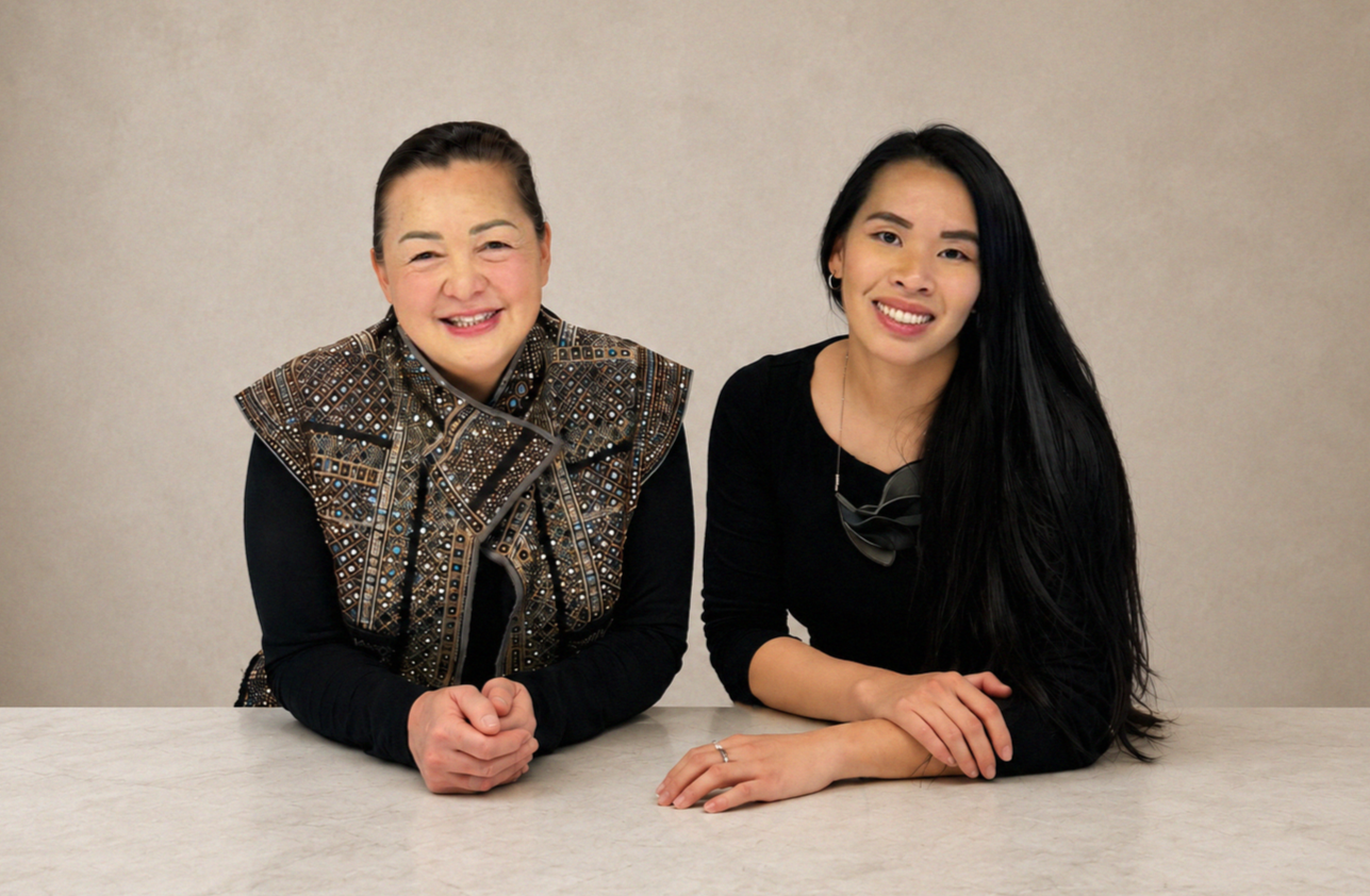 Two women sitting at a table, smiling, against a plain beige background.