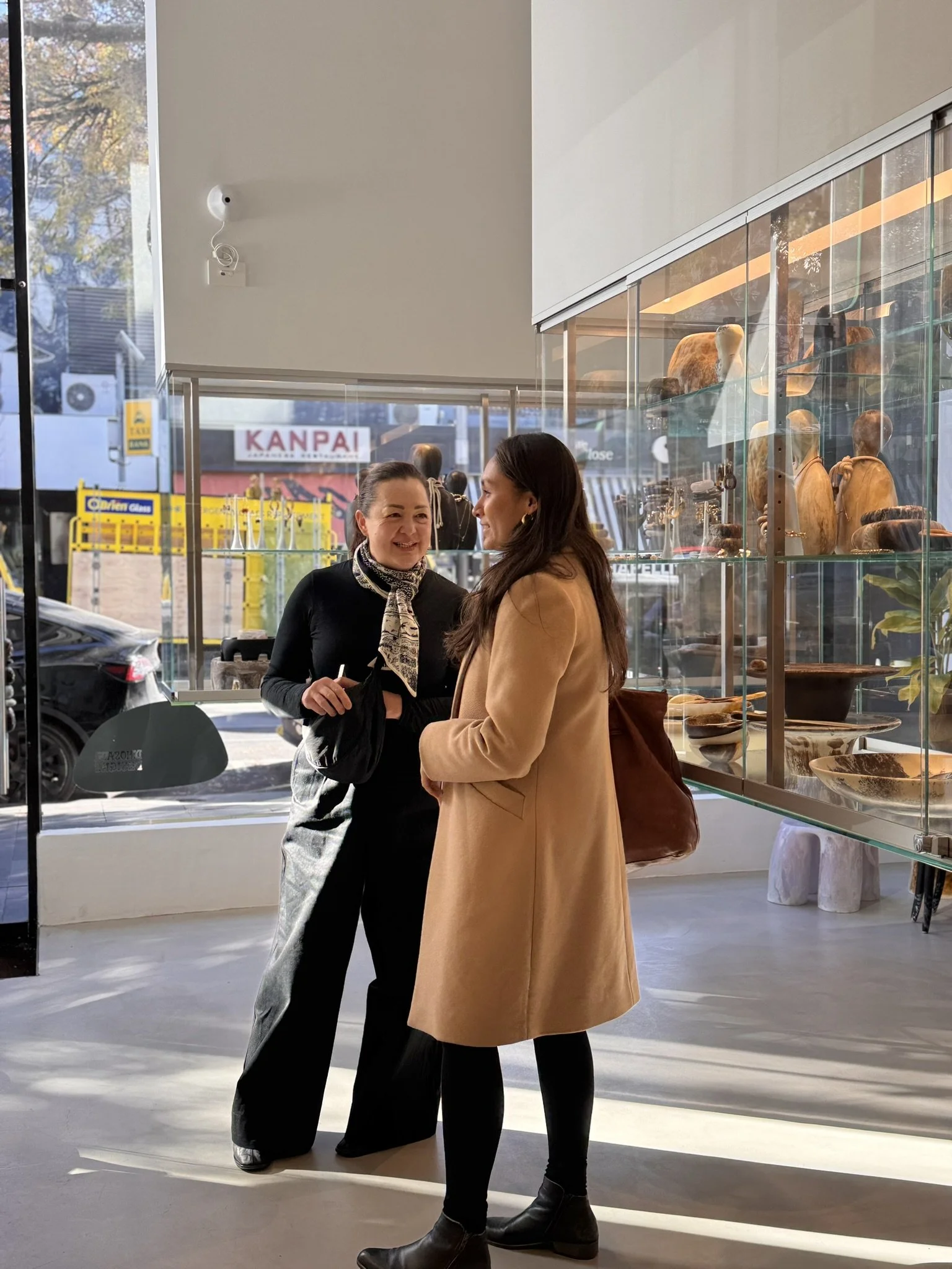 Two women are standing inside a store, having a conversation. One woman is wearing a black outfit with a patterned scarf, and the other woman is dressed in a tan coat with a brown bag. The store has display shelves with wooden sculptures and glassware, and large windows showing a street outside.