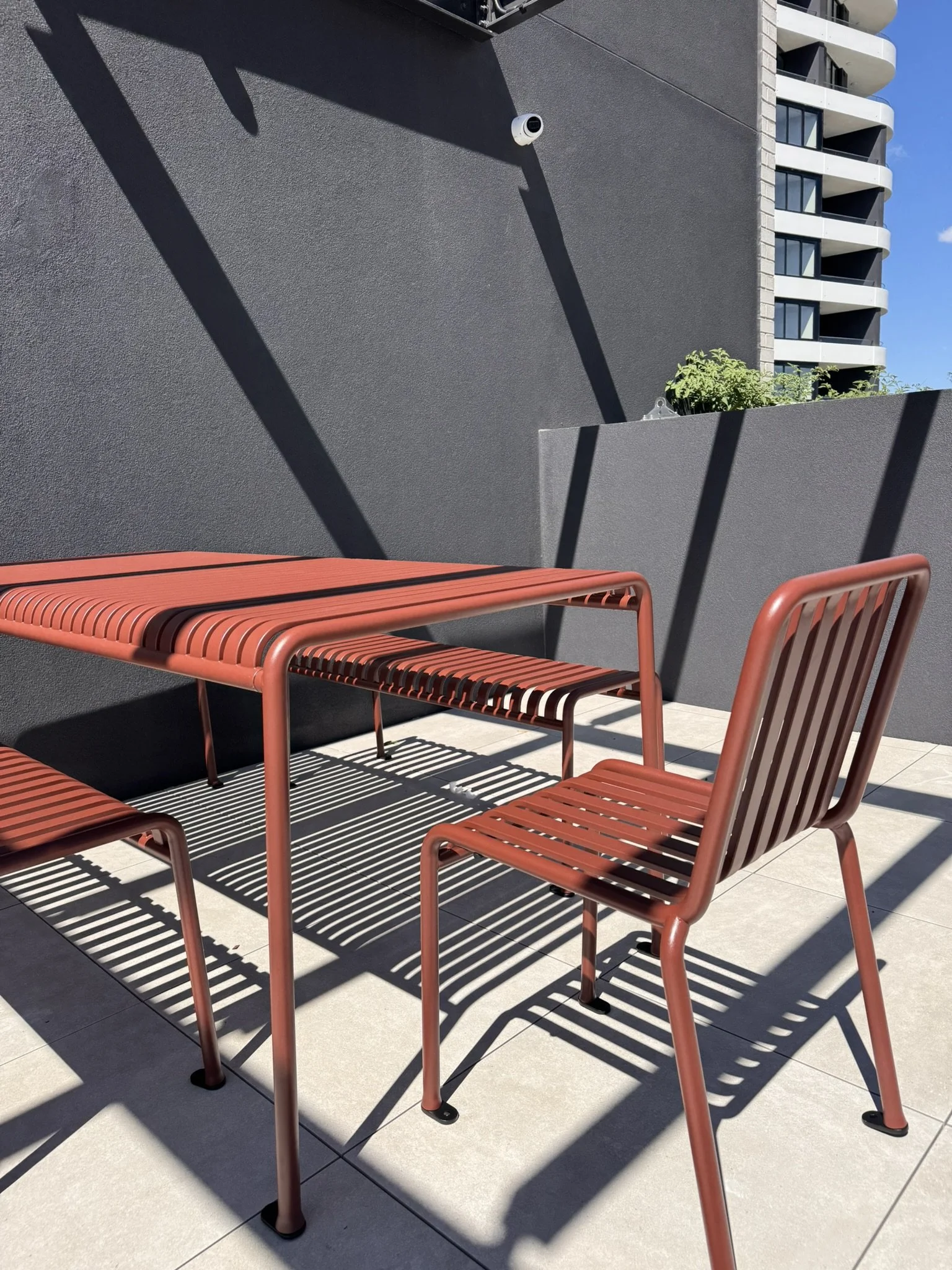 Red outdoor table with matching chairs on a tiled balcony. Shadows of the furniture are cast onto the tiles. The background includes dark gray walls with a security camera and a tall modern apartment building.