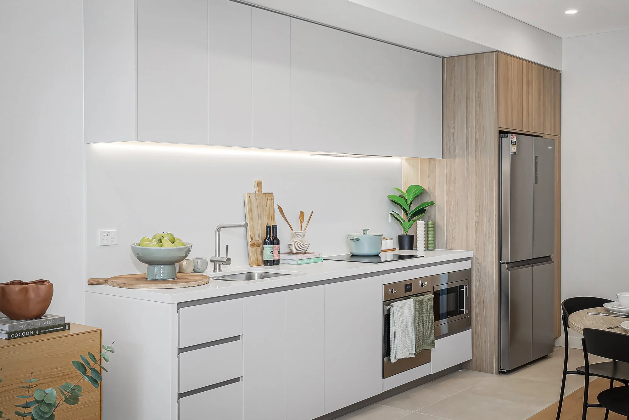 Modern kitchen with white cabinets, wooden accents, stainless steel refrigerator, and decorative plants.