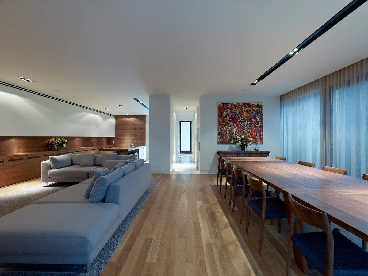 Open-concept living and dining area with wooden flooring, a large wooden dining table with eight chairs, a gray sofa, artwork on the wall, and large windows with sheer curtains.