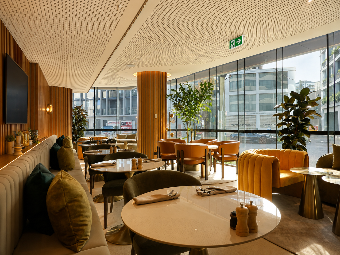 Interior of a modern restaurant with large glass windows, wooden accents, and a variety of seating including a long cushioned bench with pillows, round tables with chairs, and a cozy orange velvet armchair near a large green plant.