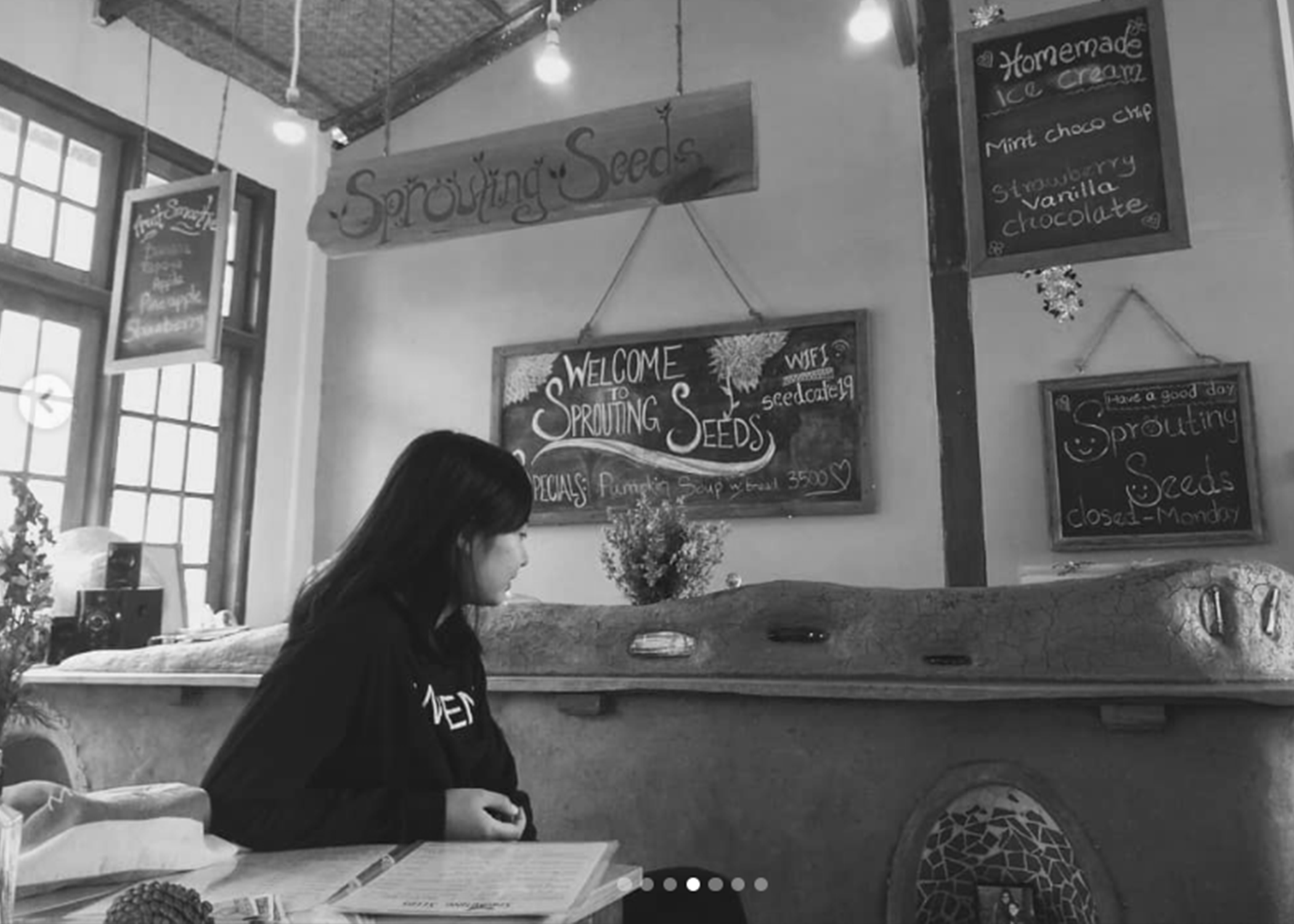 A woman sitting at a table in a cozy shop called 'Sprouting Seeds' with various chalkboard menus and signs in the background, including one advertising homemade ice cream and flavored seeds.