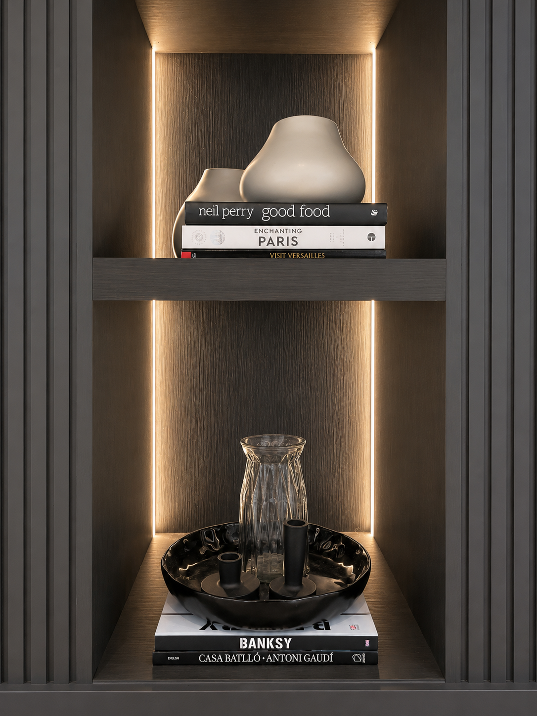 Decorative display in a dark gray cabinet with built-in lighting. The upper shelf has two white ceramic vases and three books titled 'Neil Perry Good Food,' 'Enchanted Paris,' and 'Visit Versailles.' The lower shelf features a black bowl with a textu