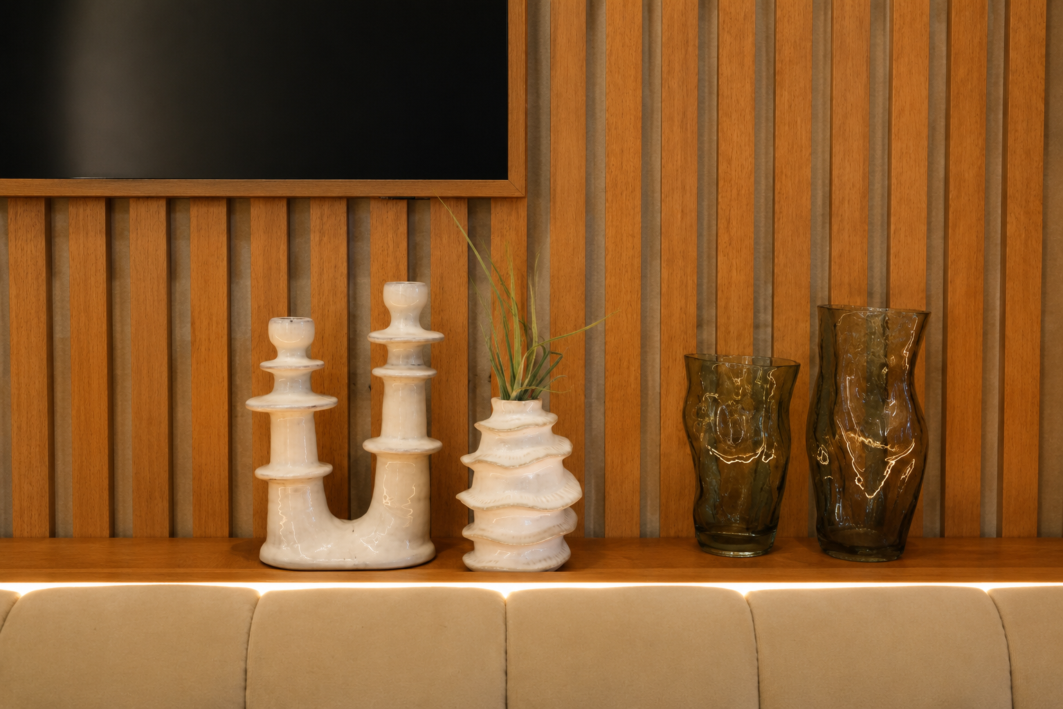 Decorative beige sofa with a wooden wall behind it, displaying four vases and a plant on a shelf. The vases include two white ceramic pieces on the left, one with a plant, and two textured translucent brown glass vases on the right.