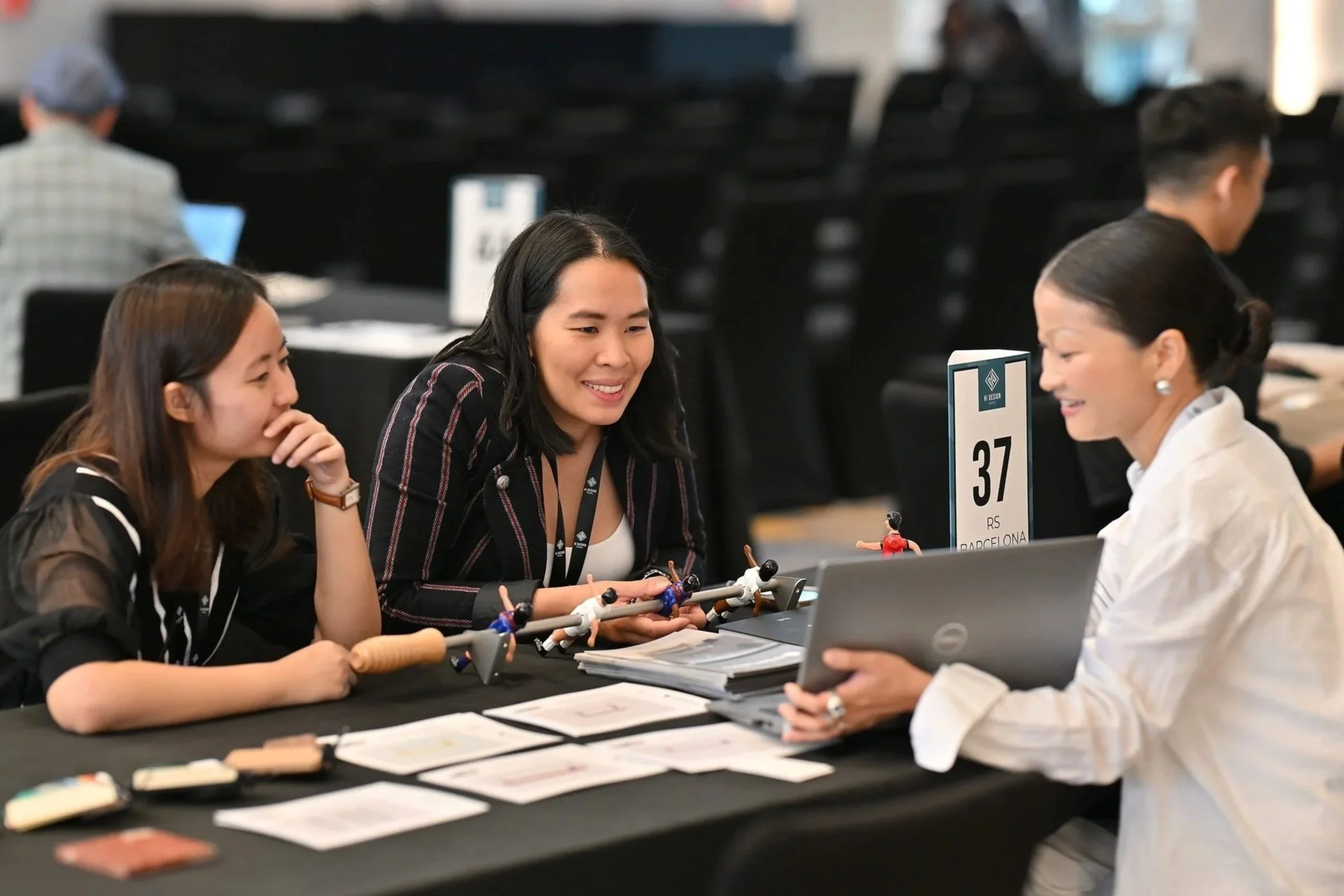 Three women sitting at a table engaged in a discussion, with a woman on the right holding a smartphone. The table has papers, pens, and miniature soccer players, and a sign indicating table number 37 from Barcelona. The background shows other tables and people in a conference setting.