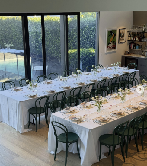 Dining room with two long tables covered in white tablecloths, set with plates, glasses, and silverware, and decorated with small flower arrangements. Large sliding glass doors reveal a view of a pool and lush greenery outside.
