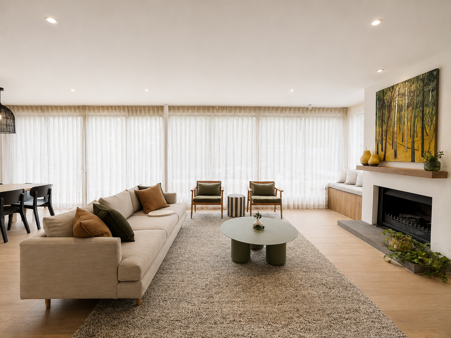 A modern living room with a beige sofa, two green armchairs, and a round coffee table on a patterned rug. The room has large windows with sheer white curtains, a fireplace with a wooden mantel, and colorful artwork on the wall. Decorative vases and p