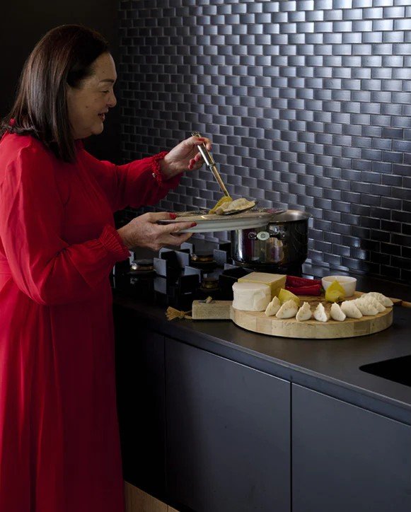 A woman in a red dress preparing cheese on a kitchen countertop with various cheeses and accompaniments, stove, and black textured backsplash.