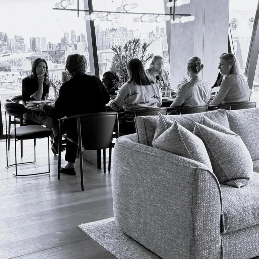 Black and white photo of a group of women sitting around a dining table with glasses of wine, in a high-rise apartment or restaurant with large windows showing a city skyline.