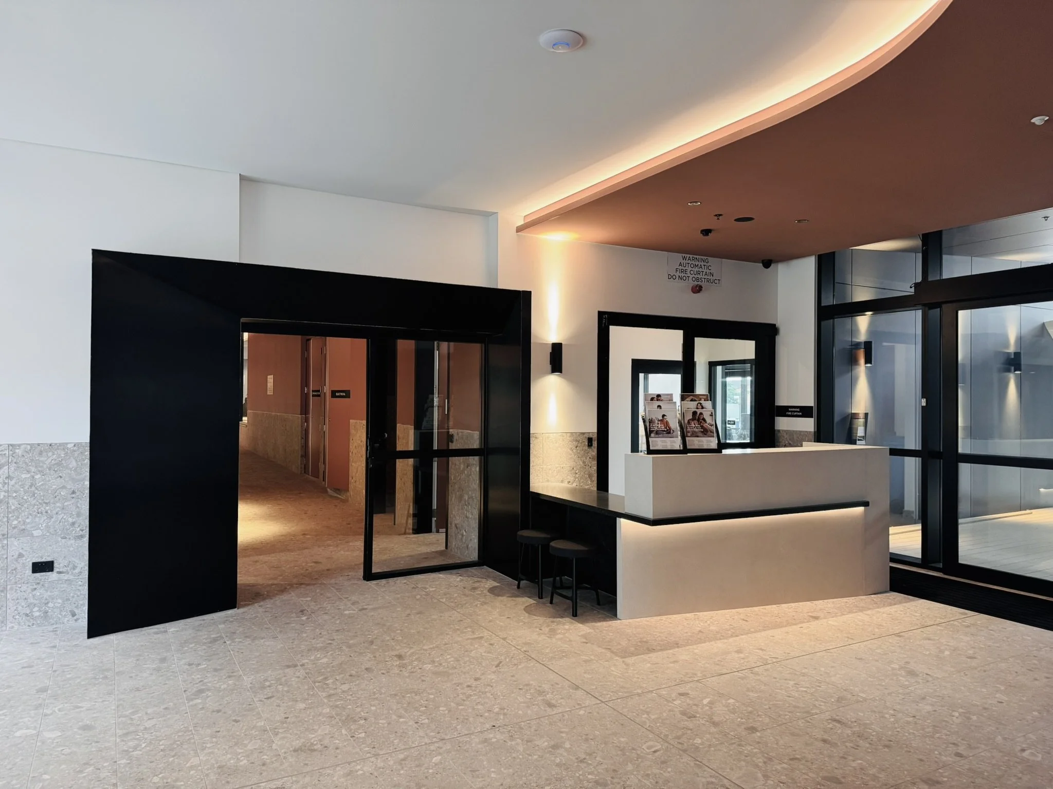 Modern hotel lobby with a reception desk, black door frame, and glass entrance doors. Light-colored flooring and walls with warm lighting.