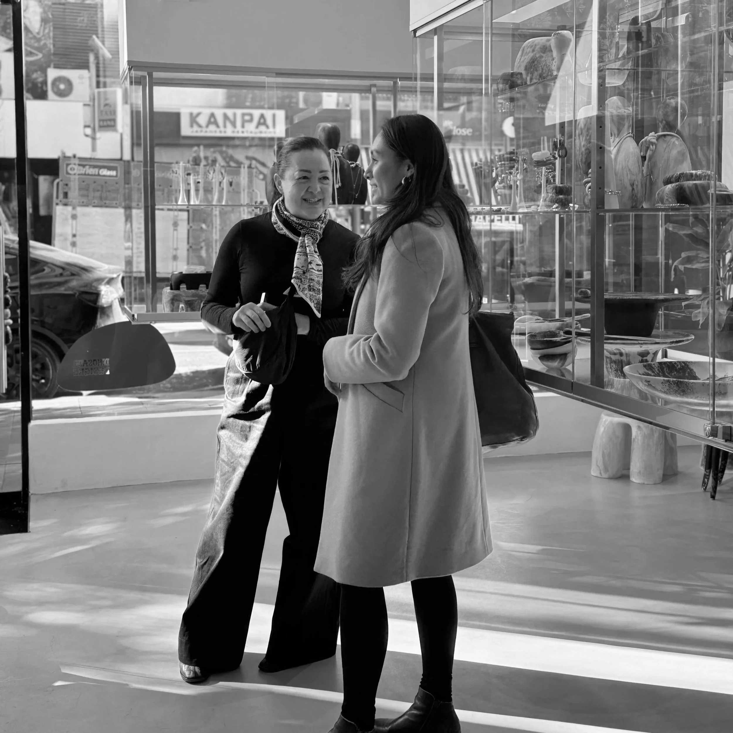 Two women standing near a glass window, engaged in conversation, with mannequins and display shelves in the background.