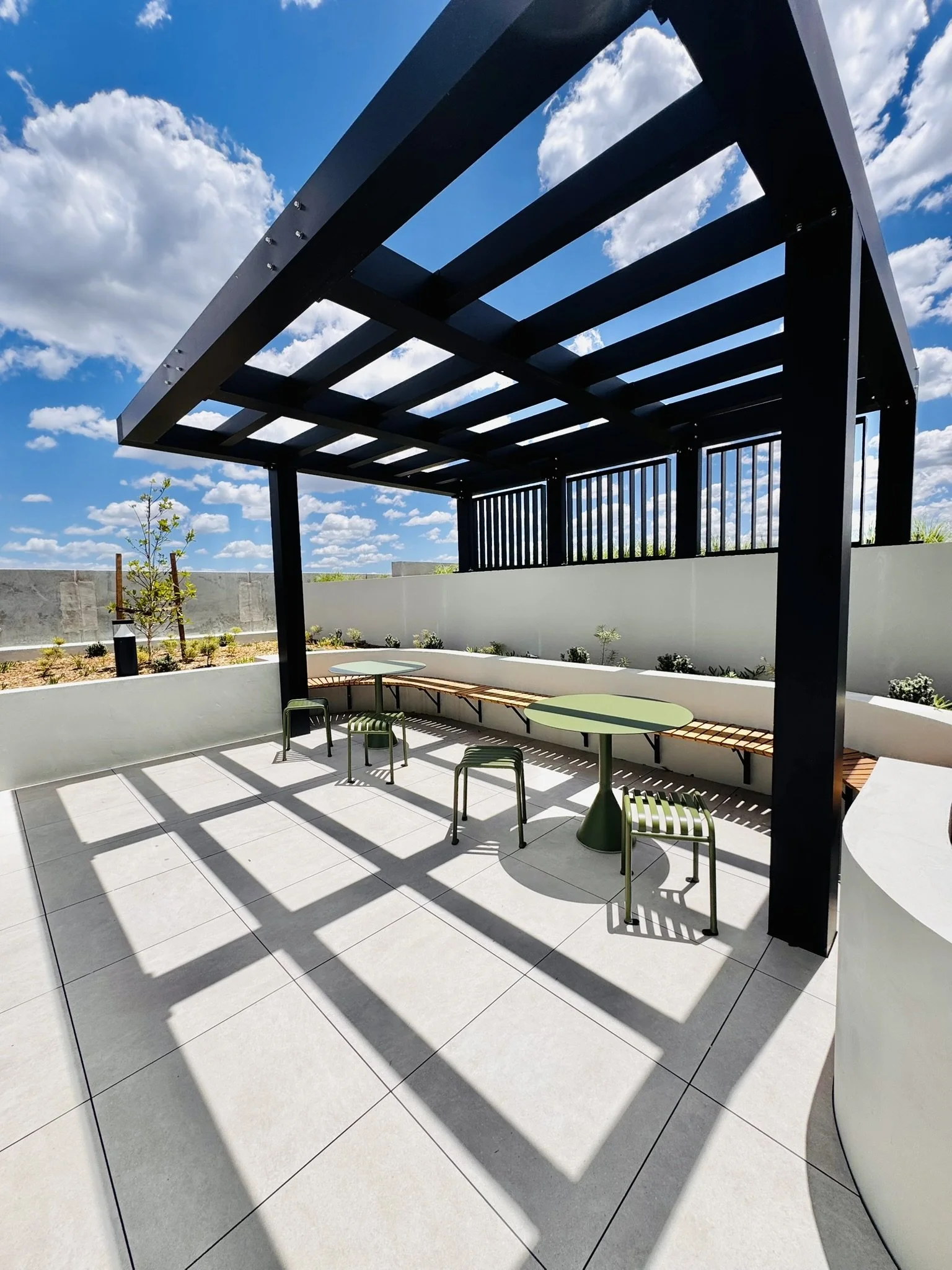 Outdoor patio with black pergola, two round green tables, four green chairs, white curved walls, and partly cloudy sky.