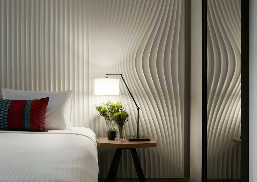 A bedroom with a white bed, a lounge chair with a colorful pillow, a side table with a plant and a lamp, and a textured wall with wavy patterns.