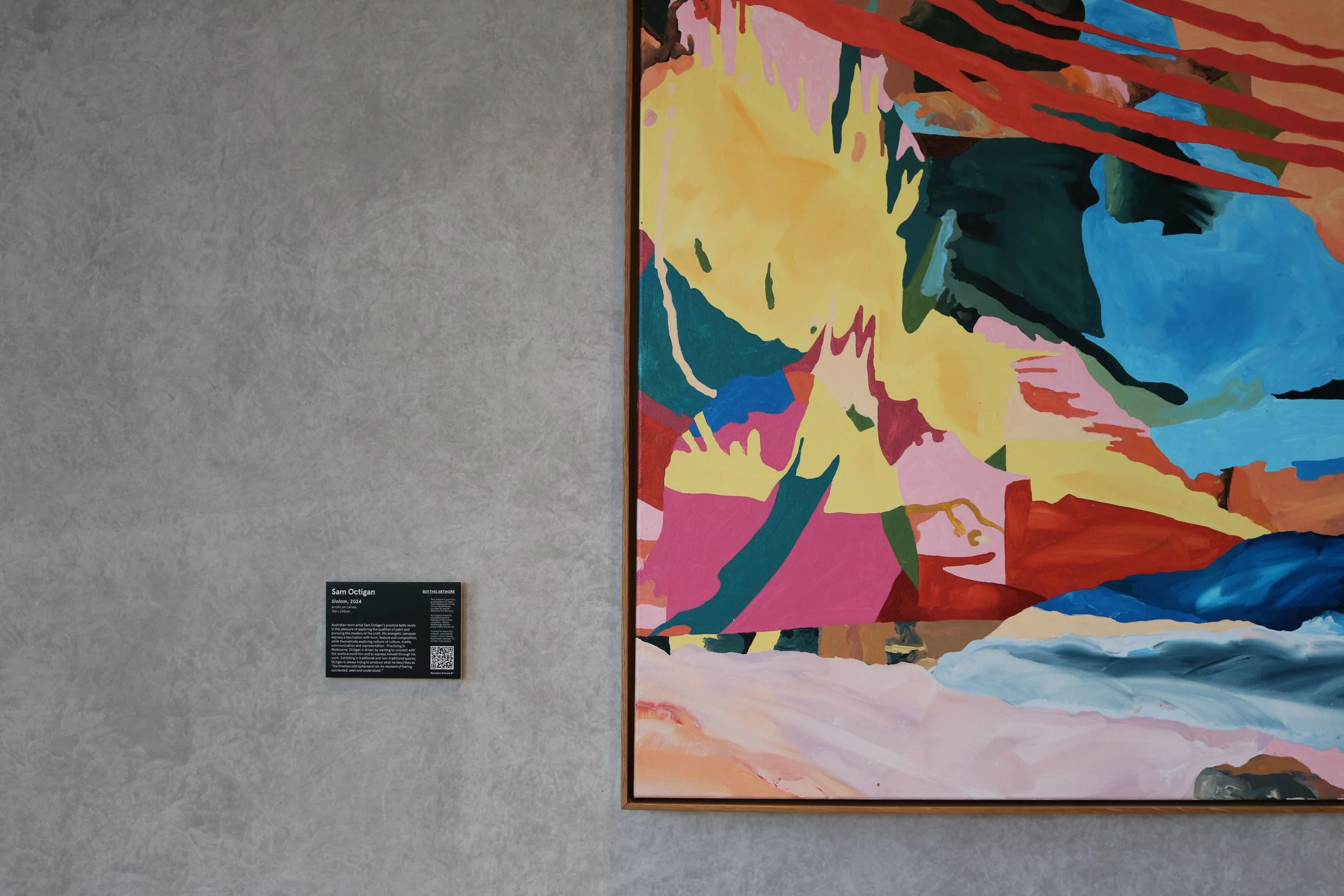 Colorful abstract painting hanging on a gray gallery wall, with a black informational sign beside it that reads 'Sam Octigan, Salom, 2024'.