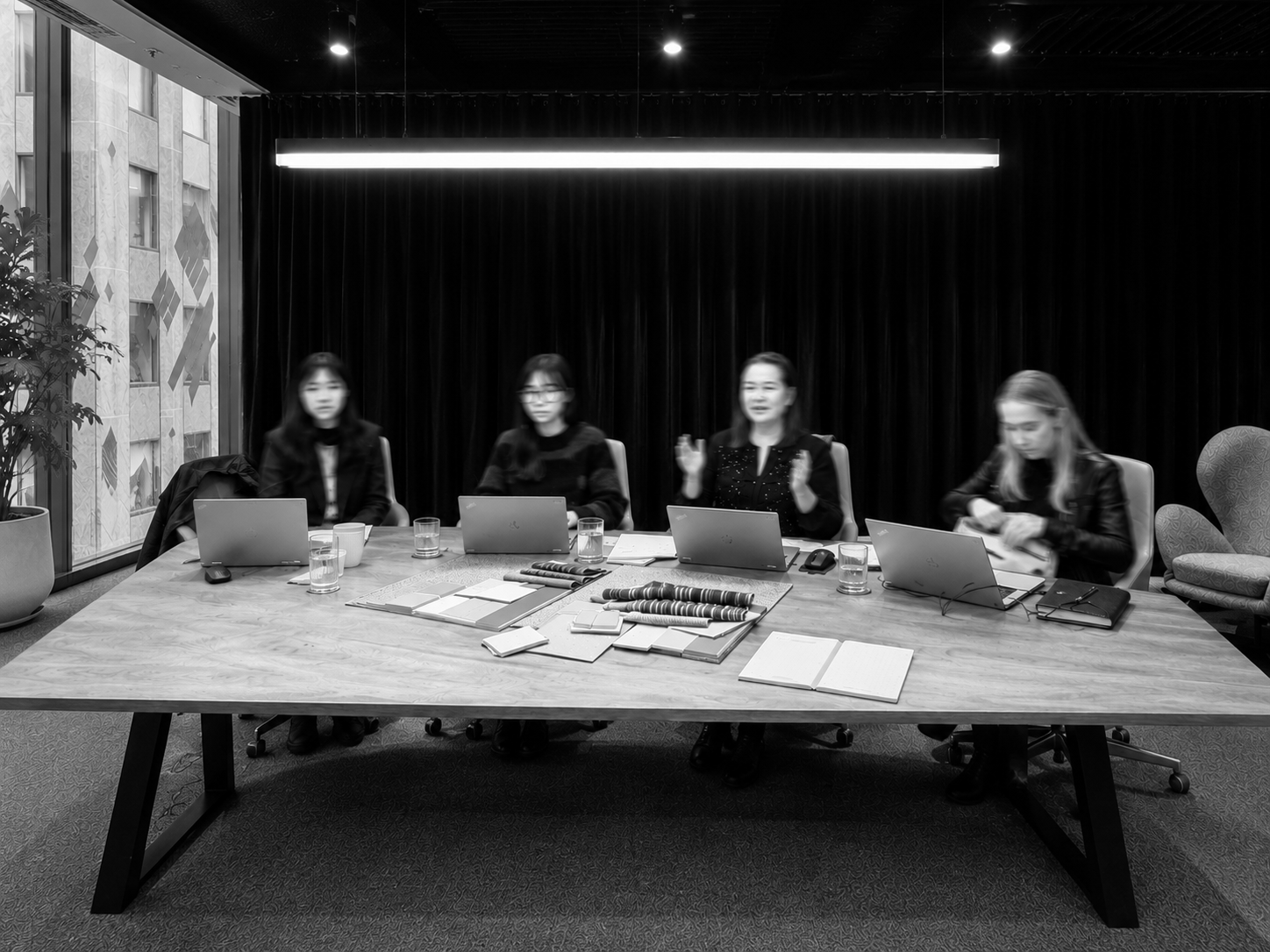 A black and white photo of a business meeting inside a modern conference room with large windows. Four women are seated at a table, some with laptops, notebooks, and glasses of water. The background shows a building with multiple windows.