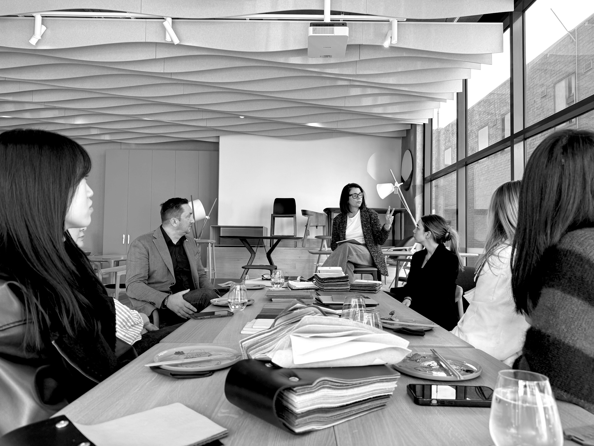 People sitting around a conference table during a meeting, with documents, notebooks, and glasses of water visible, in a modern room with large windows and a decorative ceiling.